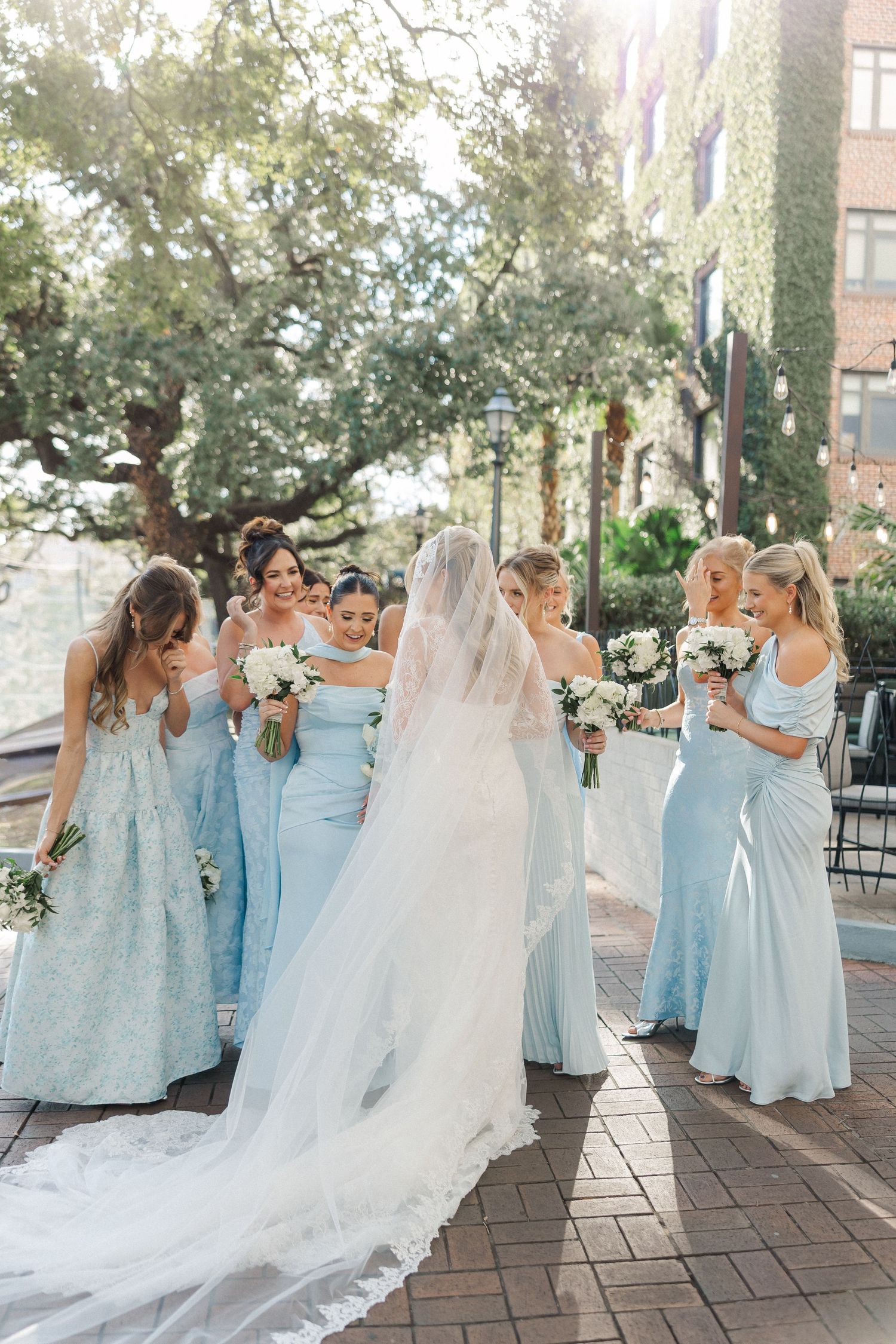 Wedding party in soft blue dresses share joyful moments on a brick patio under trees.