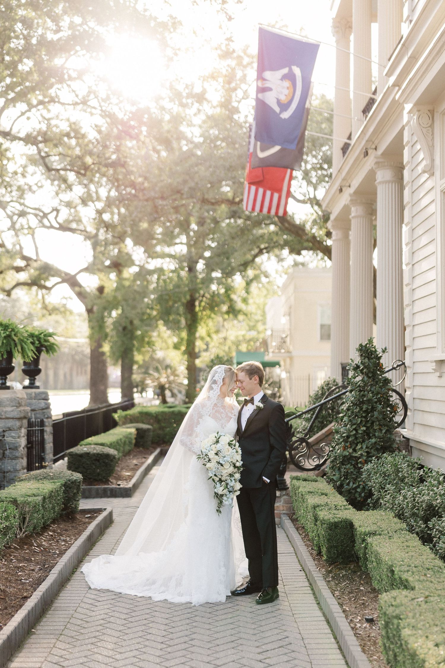 A wedding couple stands on a brick pathway outside a white columned building with a state flag hanging above.