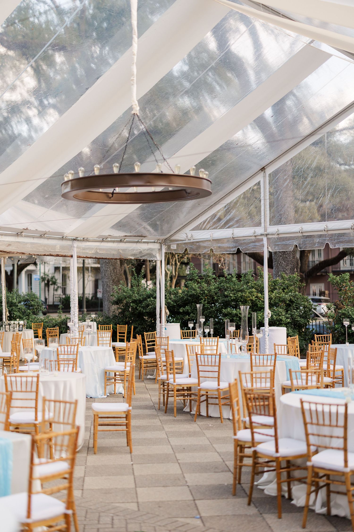 An elegant wedding reception setup with white chairs under a clear tent and hanging circular chandelier.