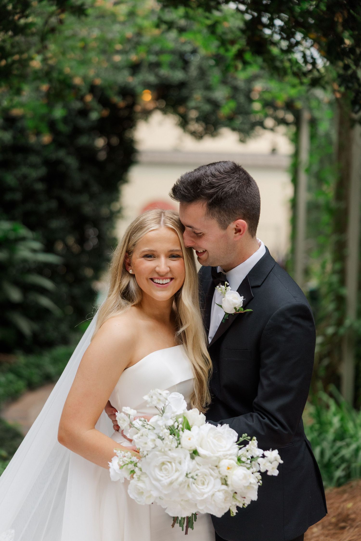 Romantic wedding couple embracing outdoors in a garden setting with white floral bouquet.