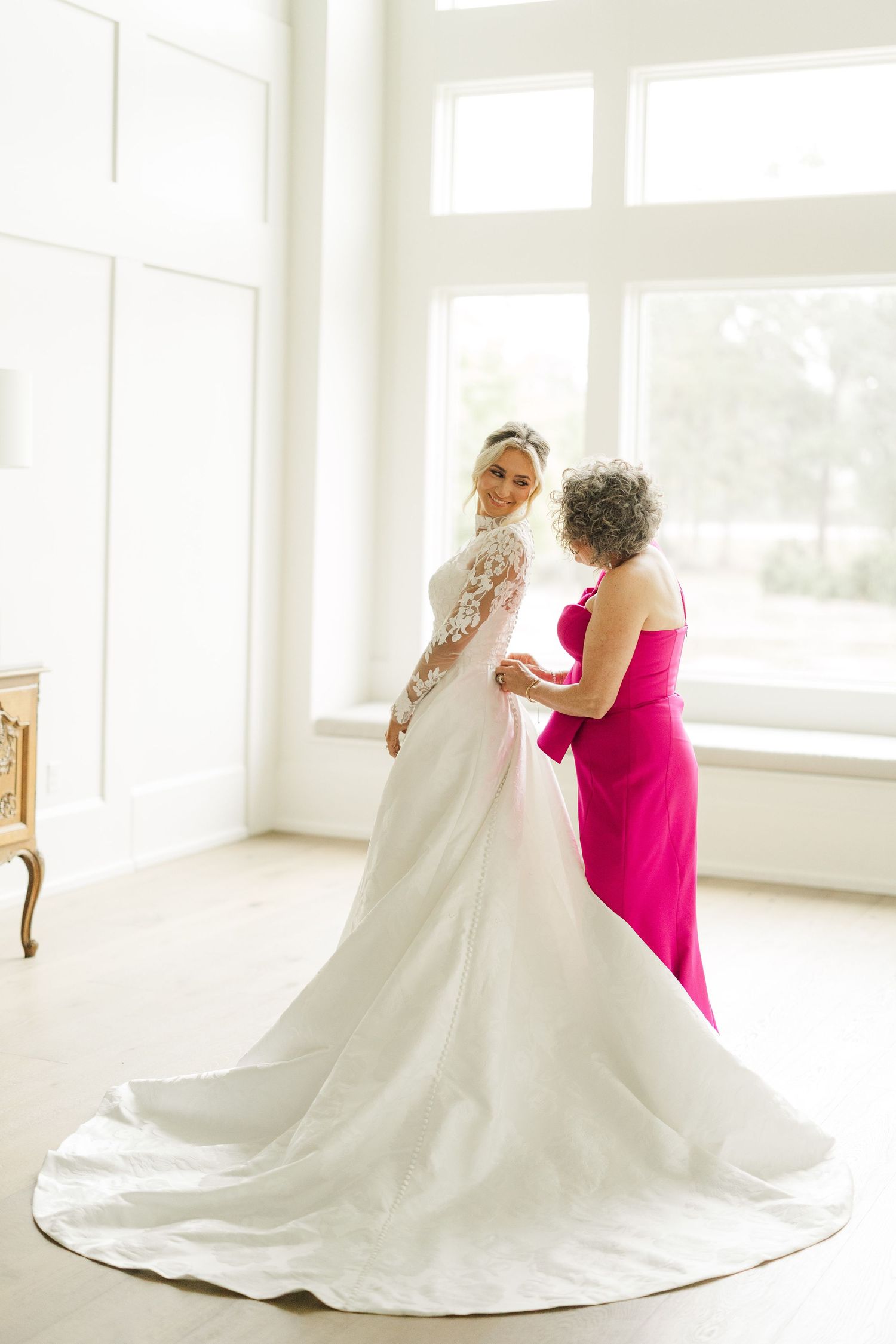 Wedding dress with long train being adjusted in bright white room with windows.