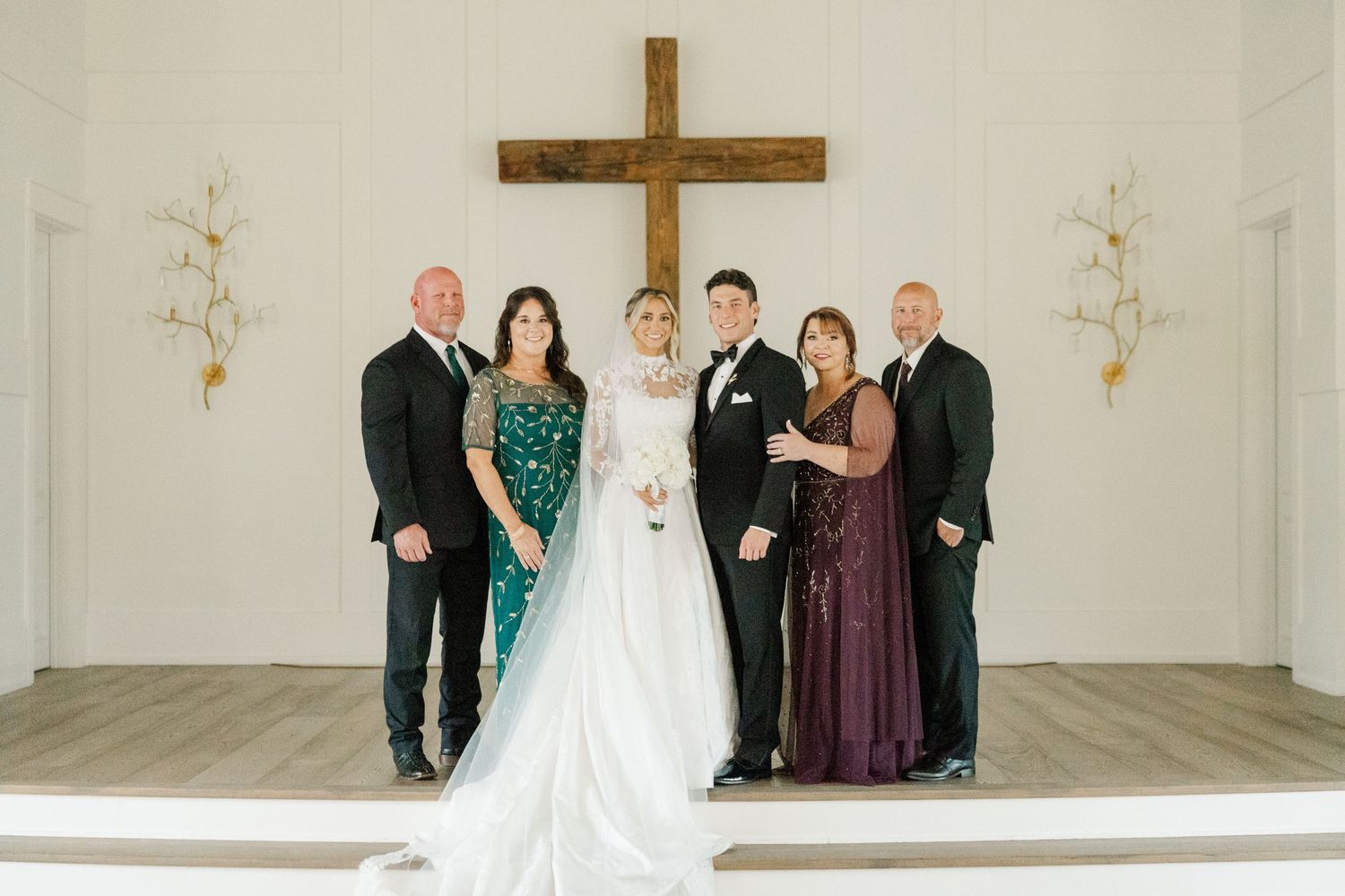 Wedding party stands in front of a wooden cross in a bright white chapel for formal photos.