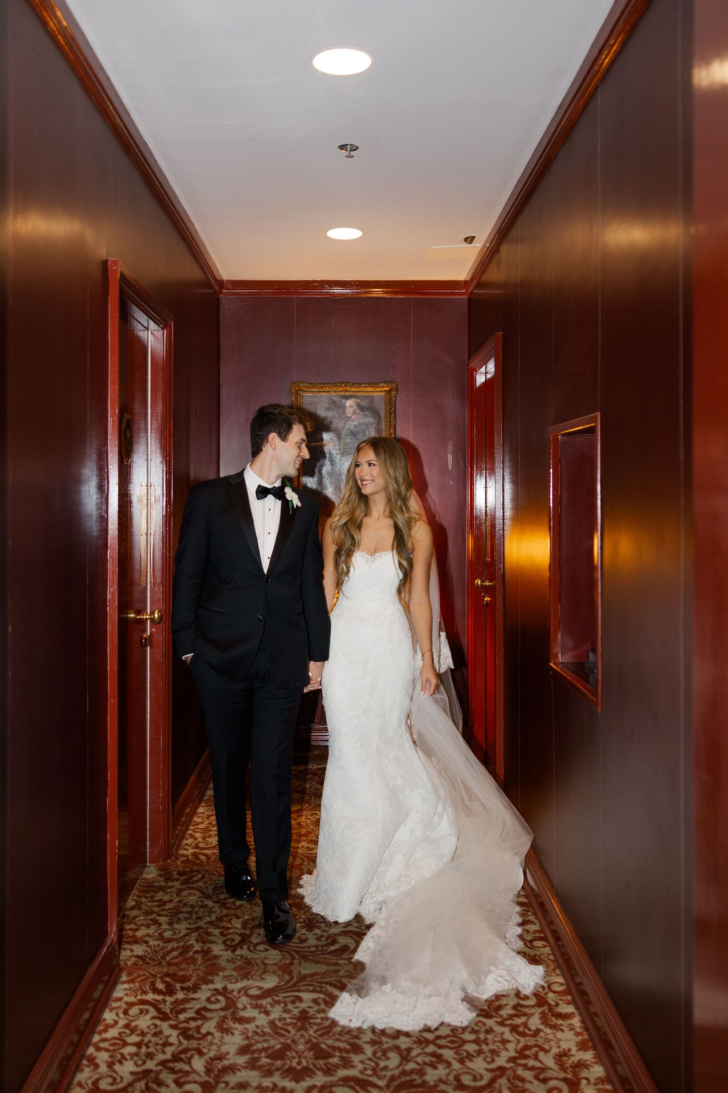 Couple in formal wedding attire walking and posing together in an elegant hotel hallway with rich wood paneling.