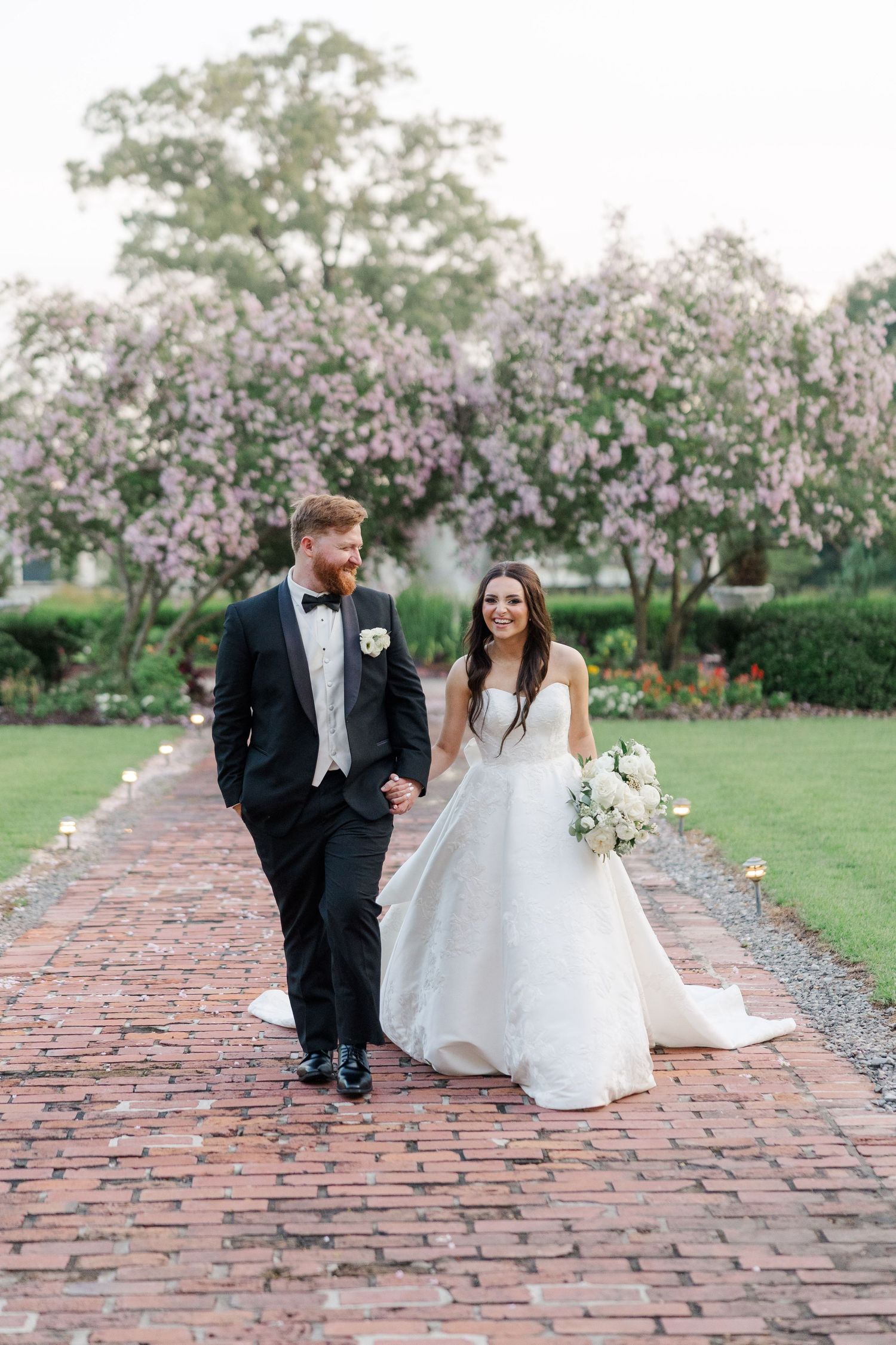 Wedding couple poses on brick pathway with blooming cherry trees in background.