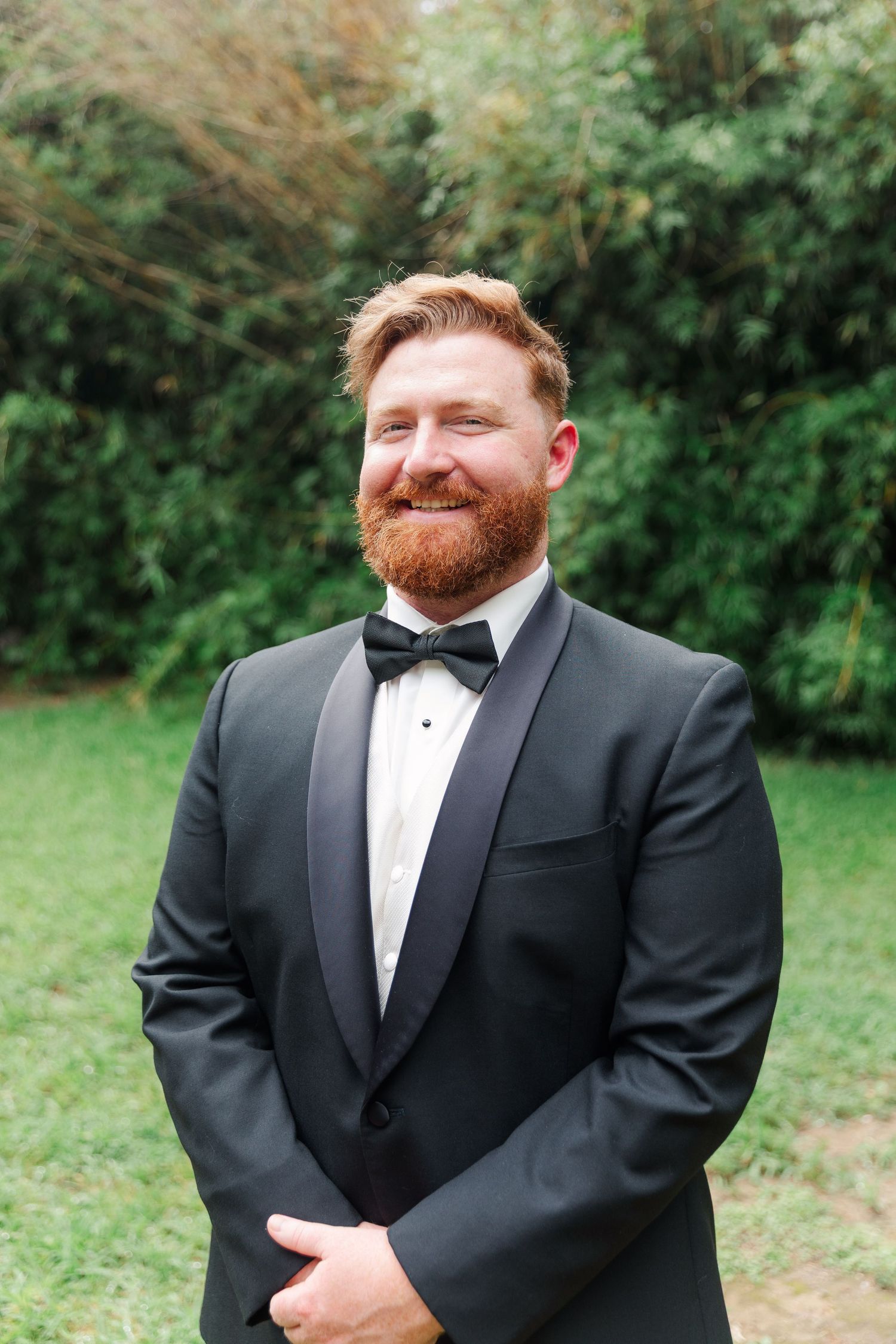 Groom in black tuxedo smiling outdoors with red beard.