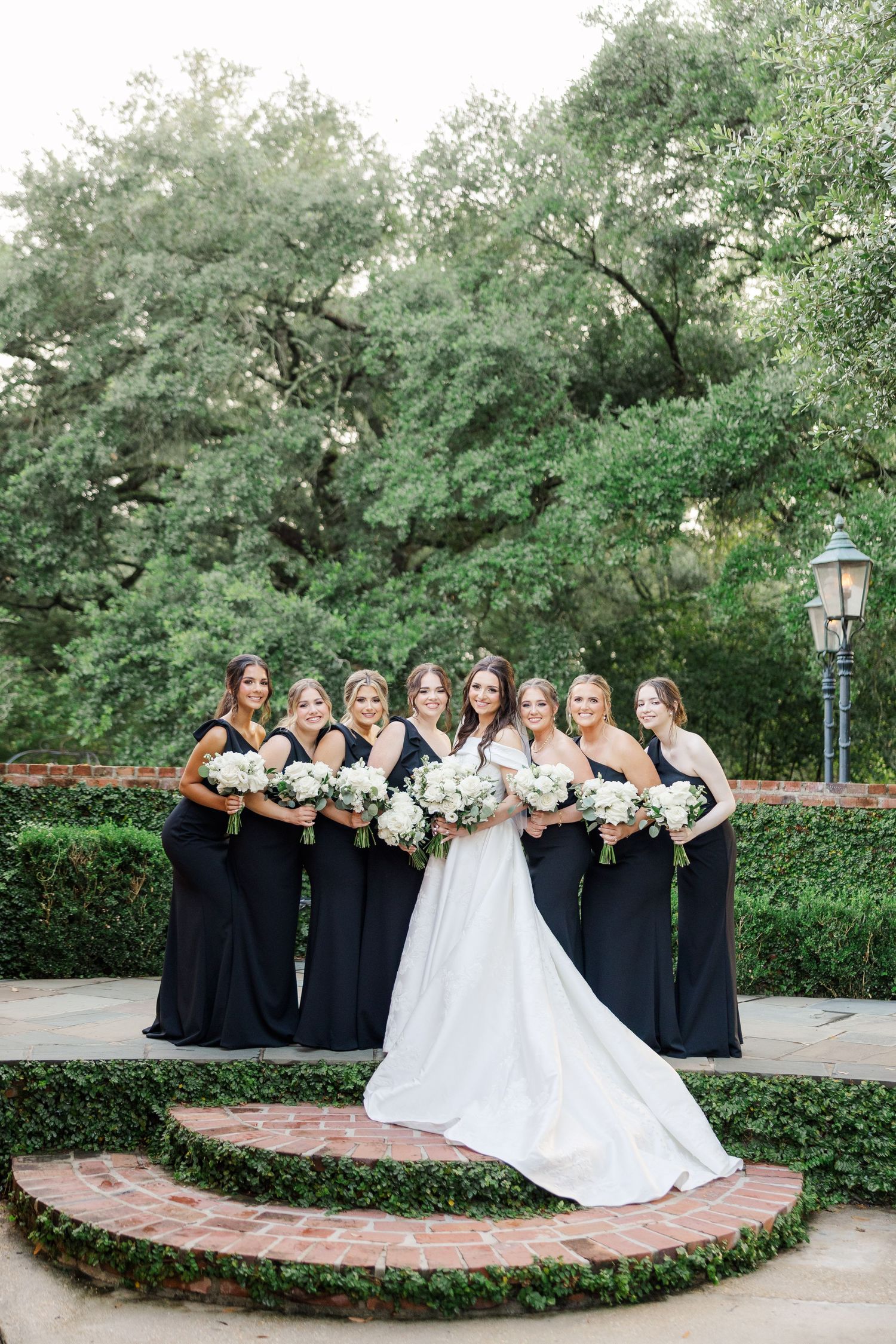Bridesmaids in black dresses pose with bride in white gown in a garden setting.