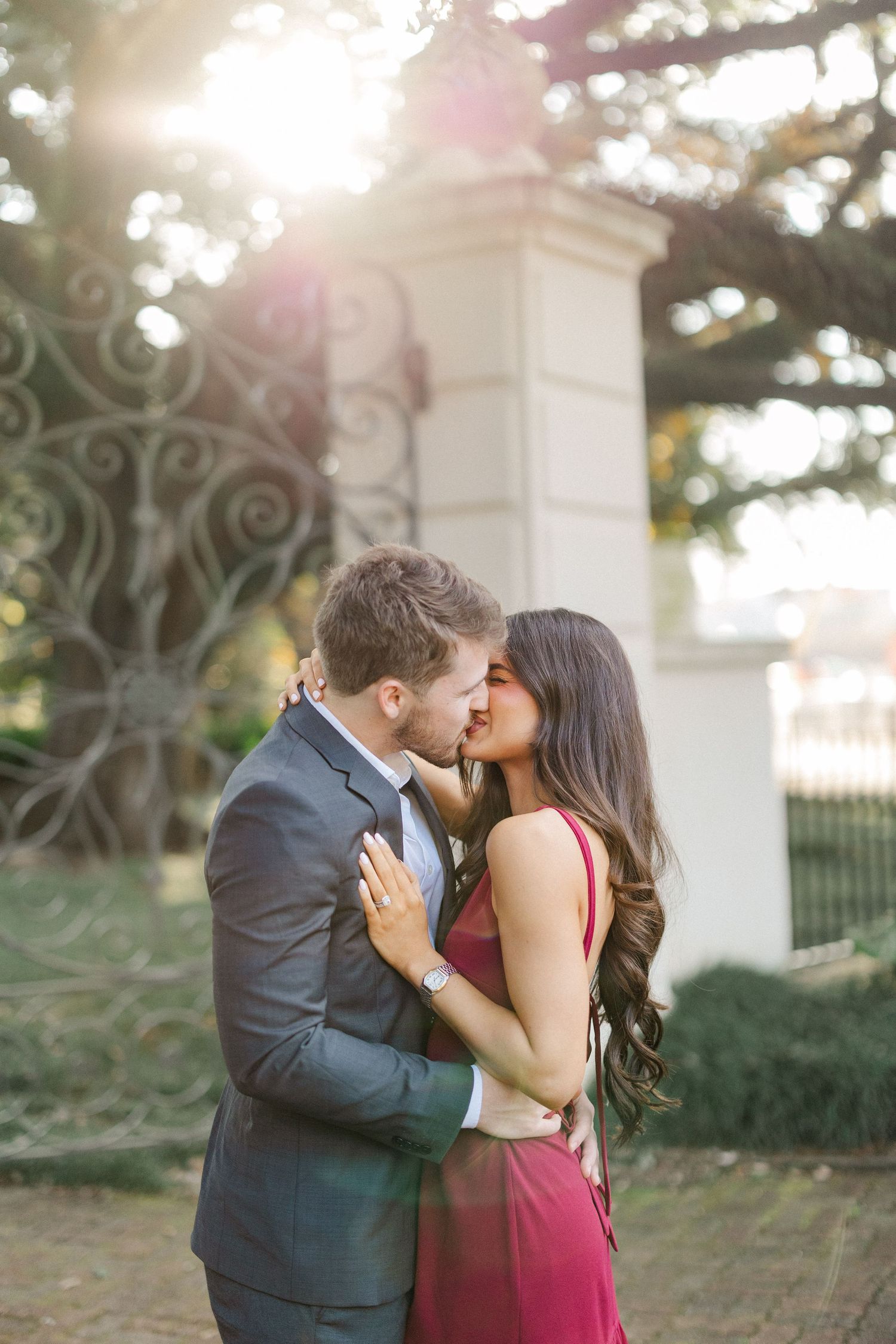 A romantic couple embraces and kisses in an outdoor garden setting with white columns and sunlit background.