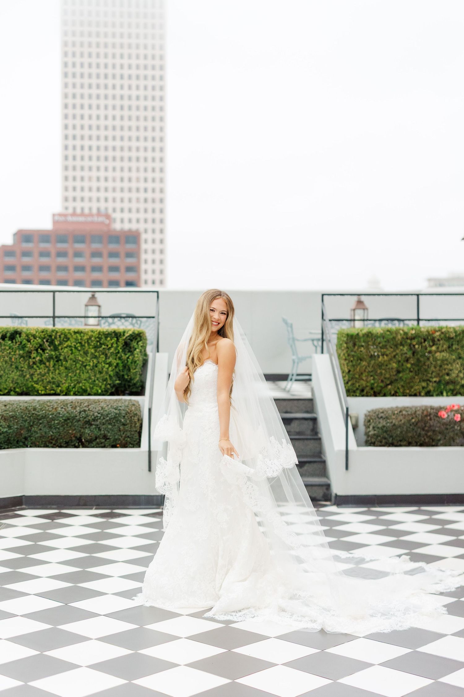 A bride in a white wedding gown poses on a rooftop terrace with a checkered floor and city backdrop.