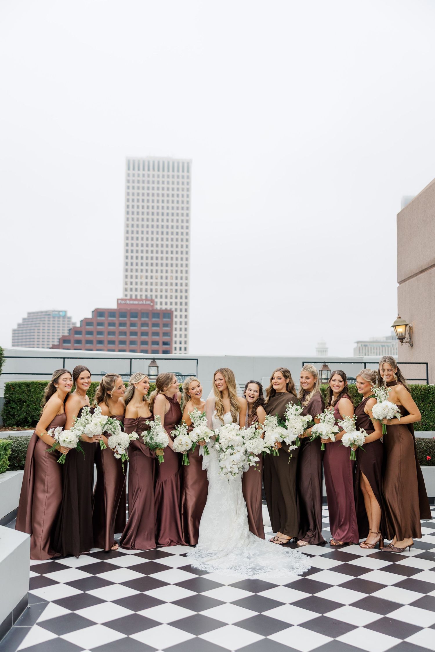 Wedding party in burgundy bridesmaid dresses poses on a rooftop with city buildings in background.