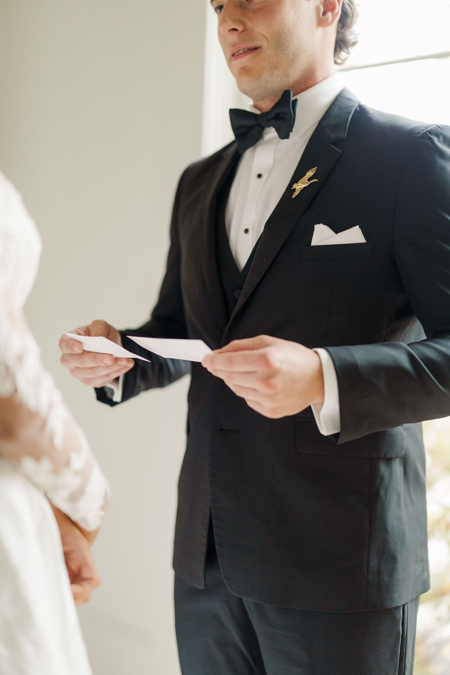 Groom in classic black tuxedo reading letter before ceremony in bright indoor setting.