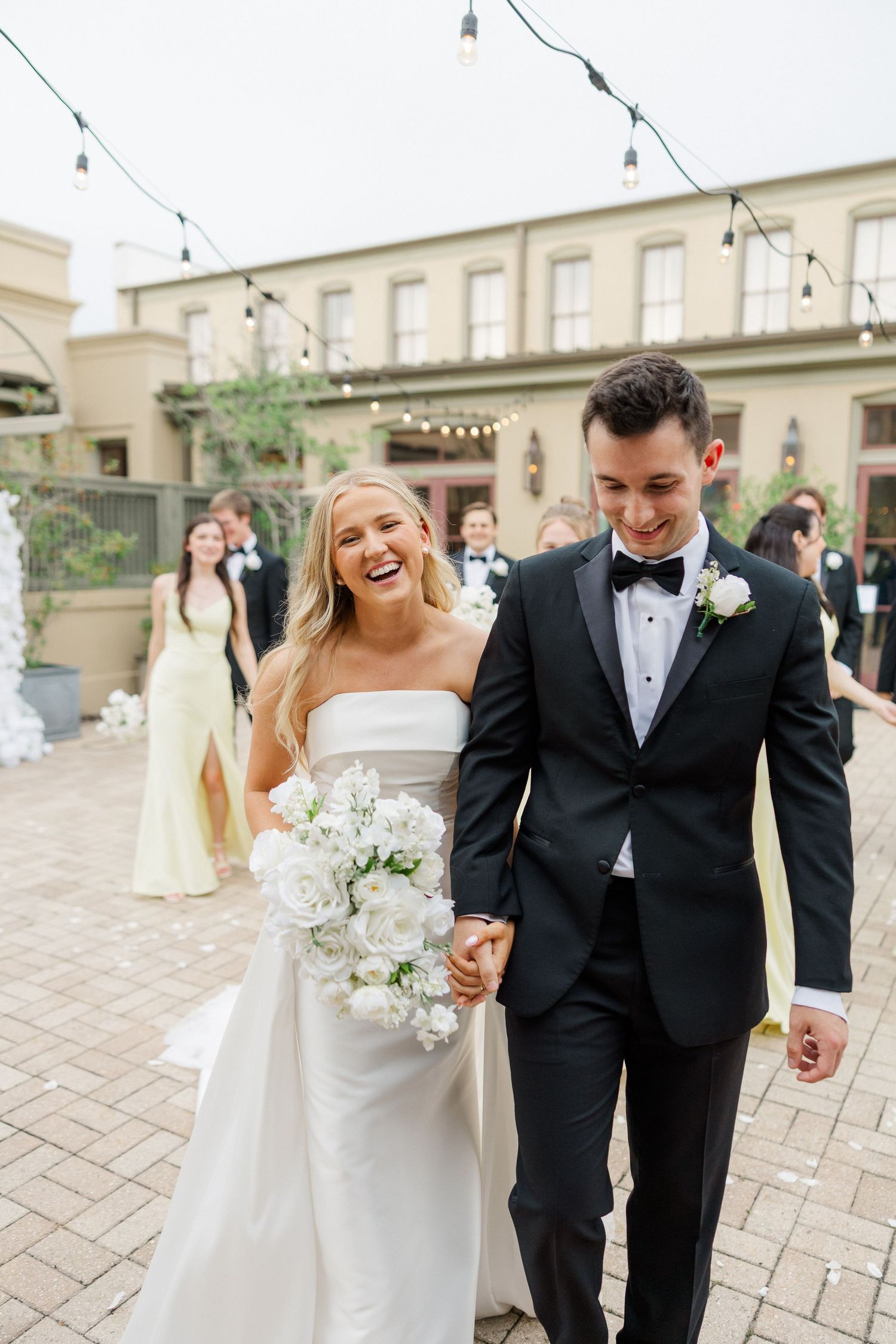 Newlyweds share a joyful moment walking together on a paved courtyard under string lights.