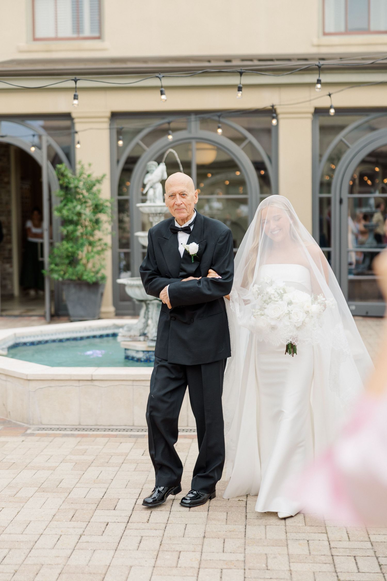 Bride in flowing white gown meeting someone on an elegant stone patio.
