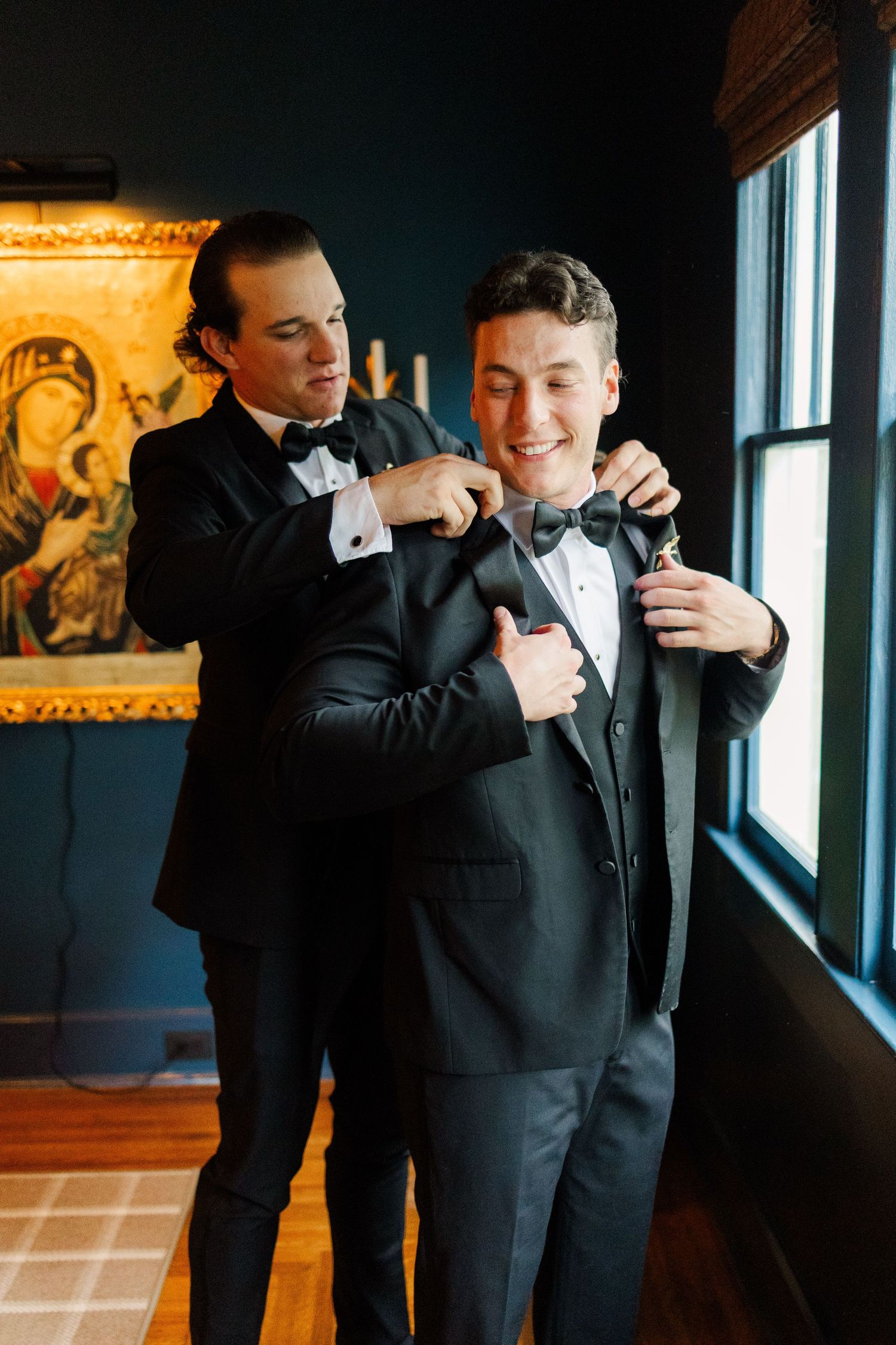 Two people in black tuxedos help each other adjust their bow ties near a window in a dark room with elegant decor.