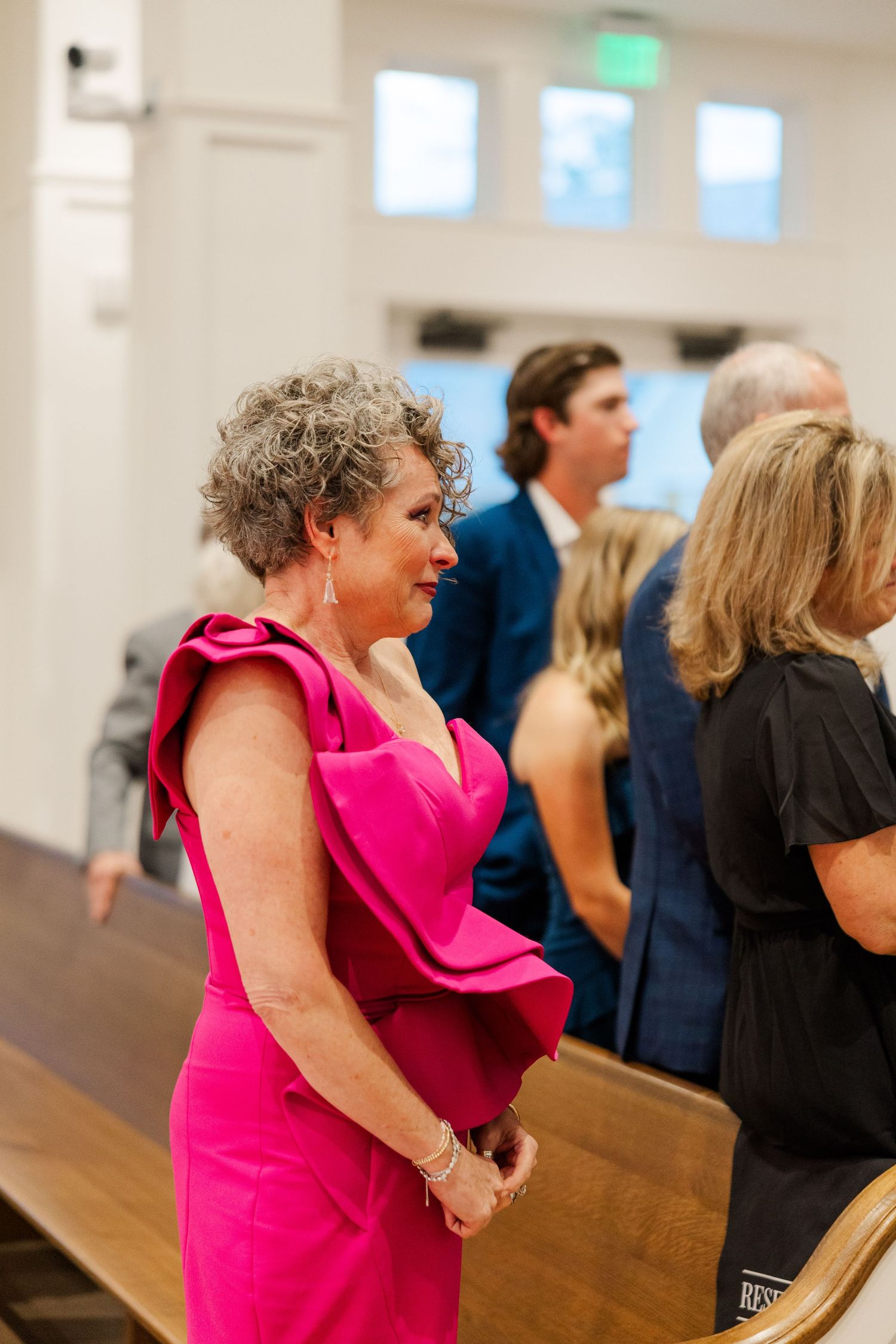 A guest in a vibrant pink formal dress stands in a church pew during a wedding ceremony.