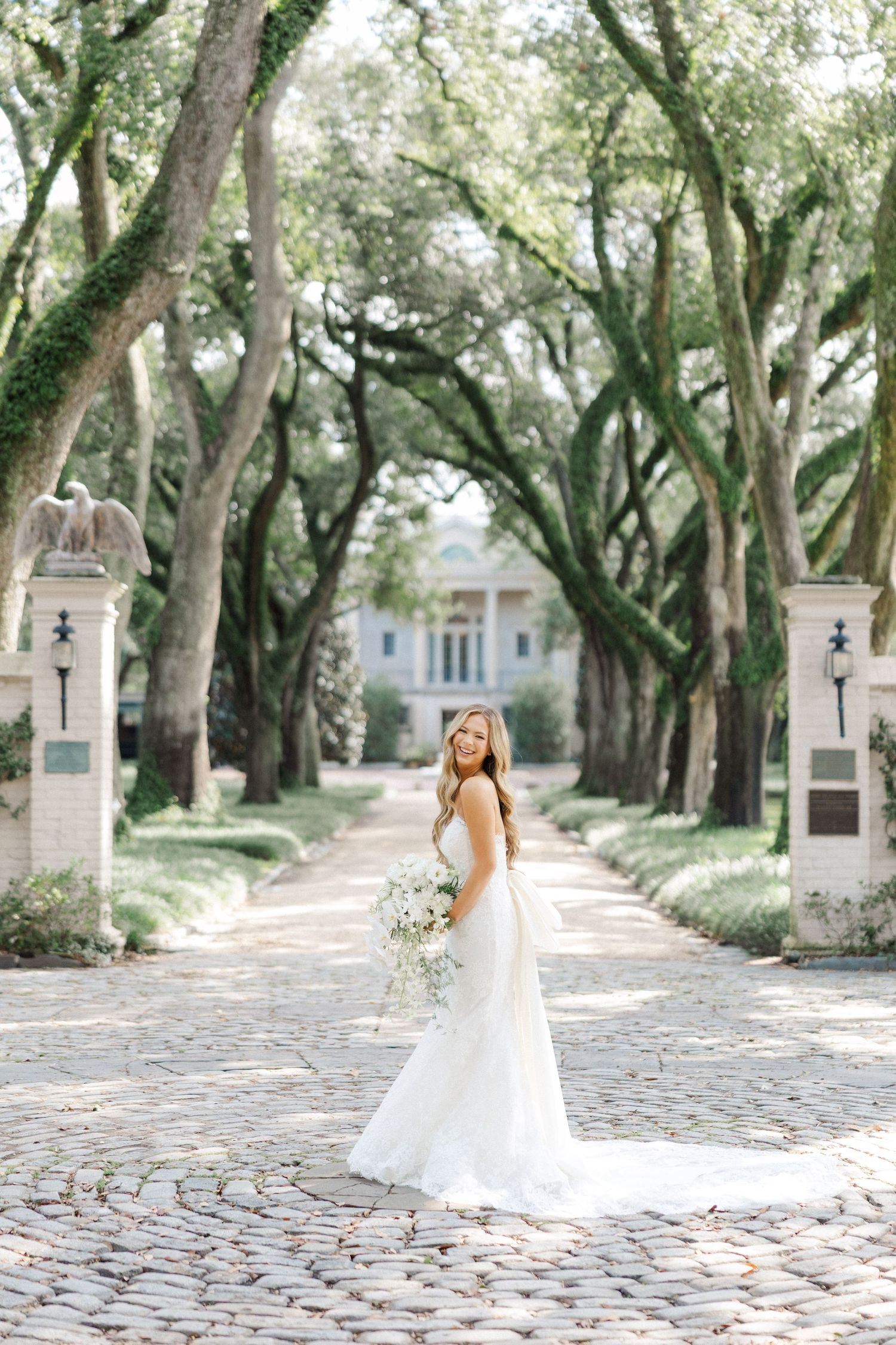 A bride in a white mermaid gown stands on a cobblestone path lined with majestic oak trees leading to an elegant mansion.