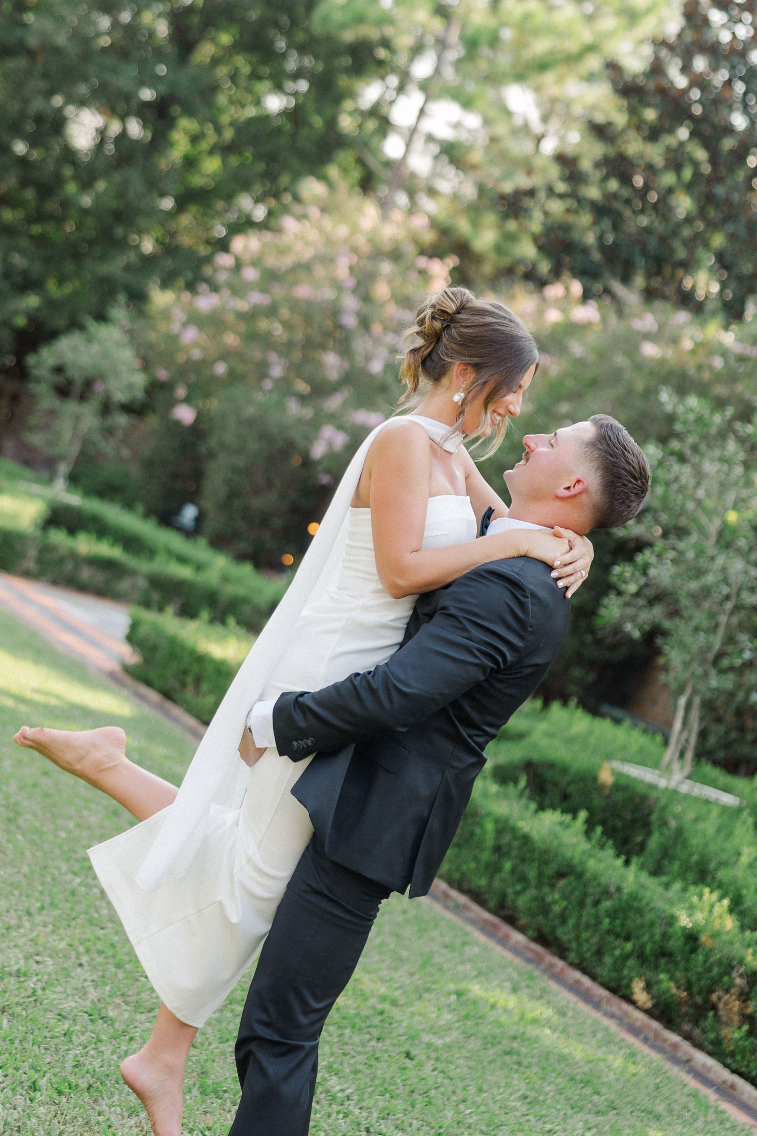 A romantic couple in wedding attire share joyful embraces and playful lifts in a lush garden setting.