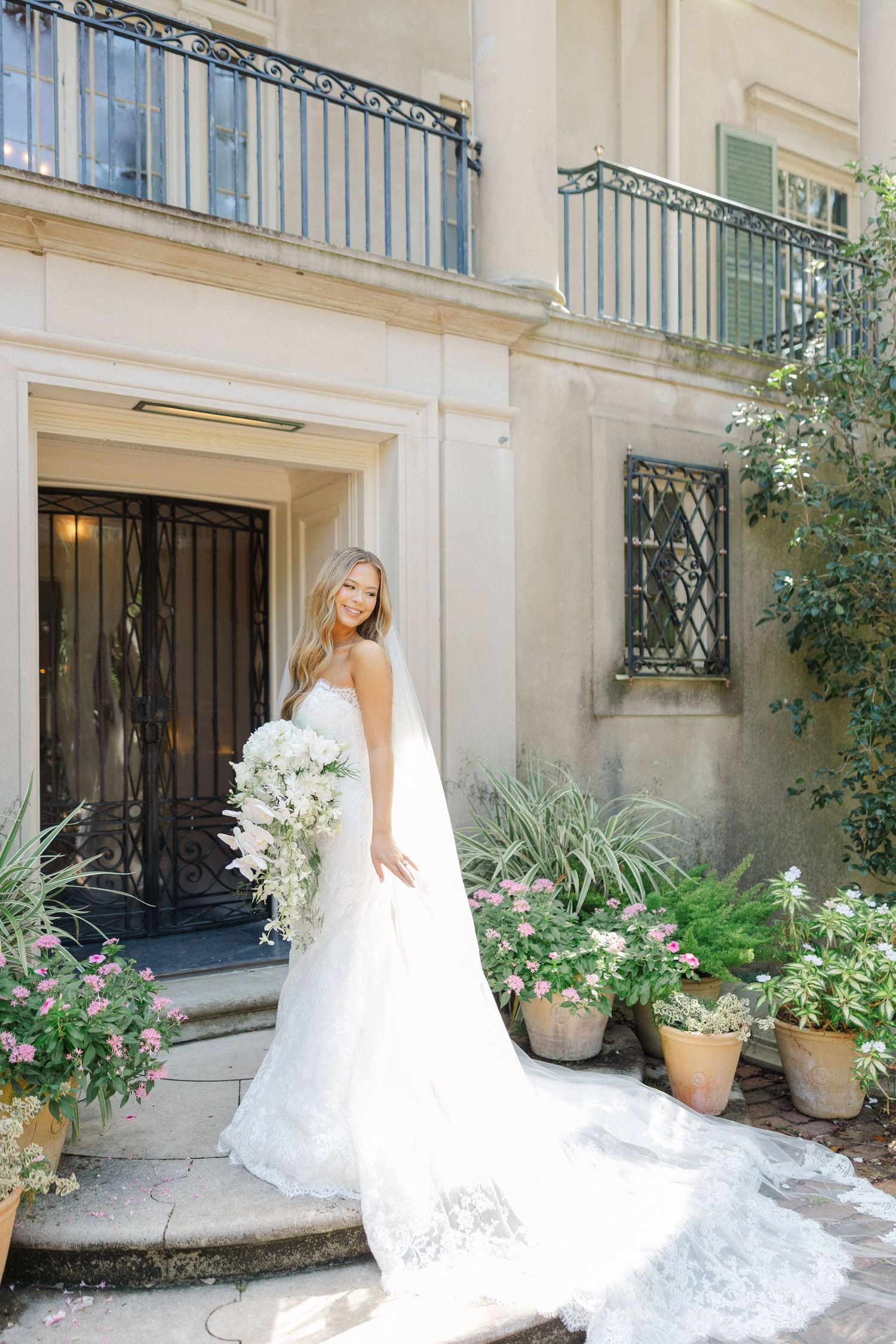A bride in a flowing lace gown twirls her dress in front of a garden setting with potted plants and iron railings.