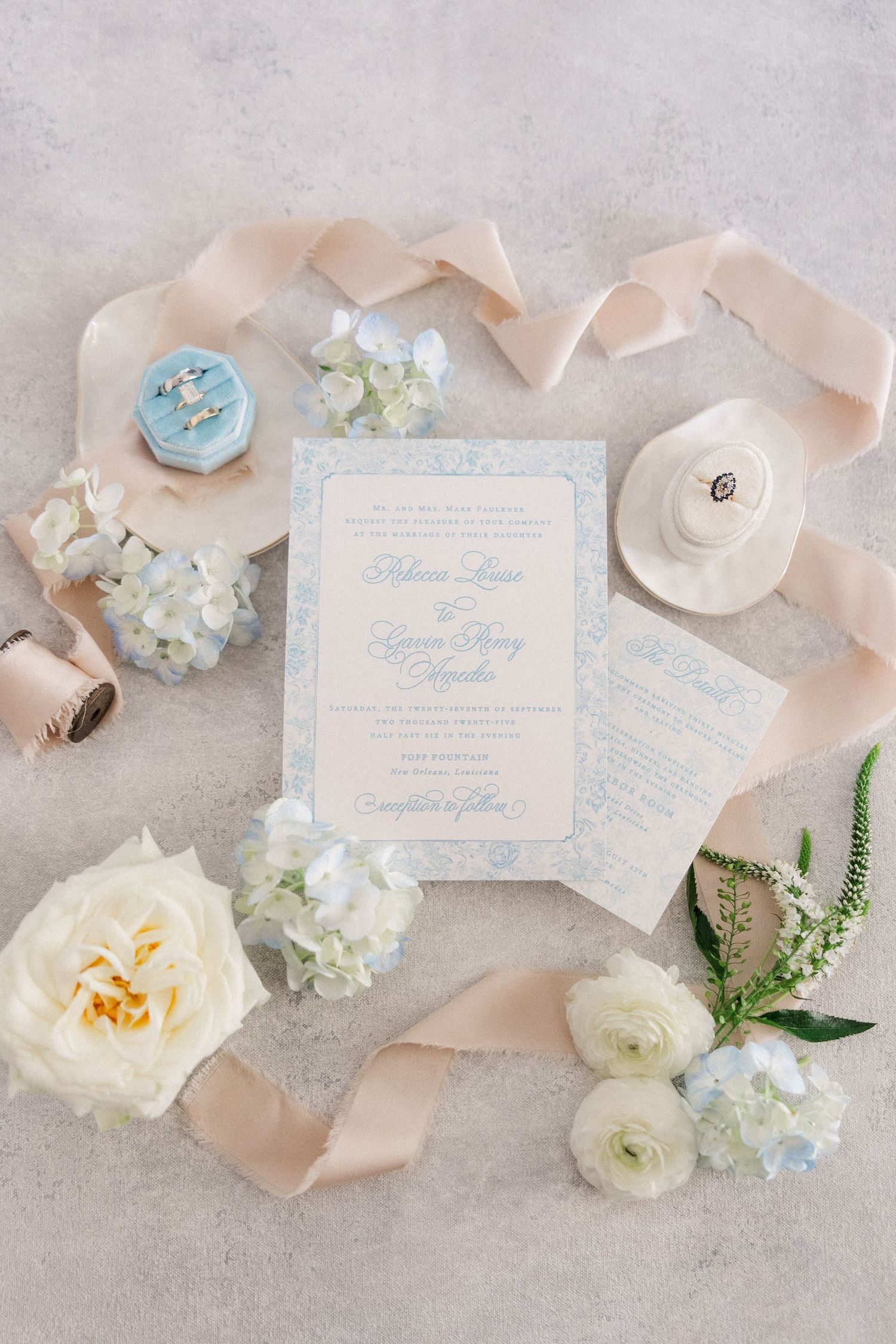 Elegant wedding invitations with blue calligraphy text displayed with white flowers and peach ribbon on grey background.