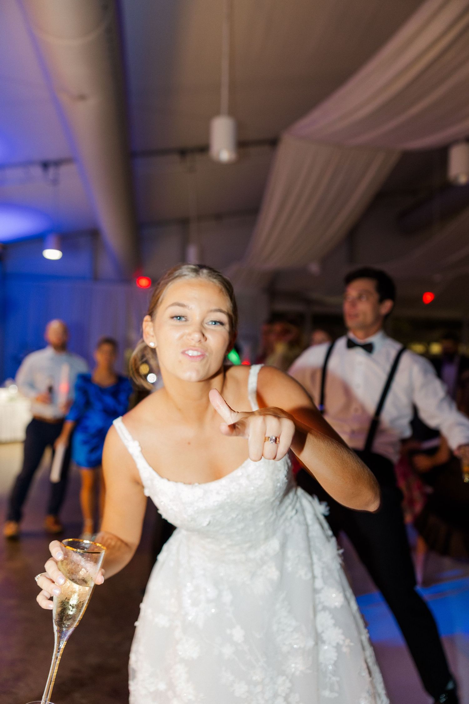 A bride in a white lace dress dances and celebrates at her wedding reception with a champagne glass.