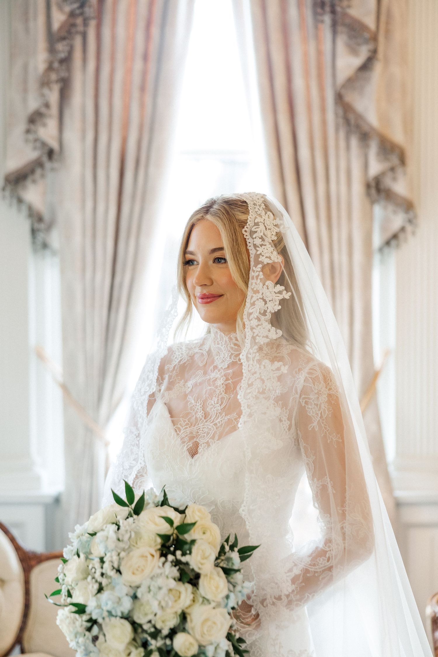 Bridal portrait with lace veil and white rose bouquet in elegant interior.