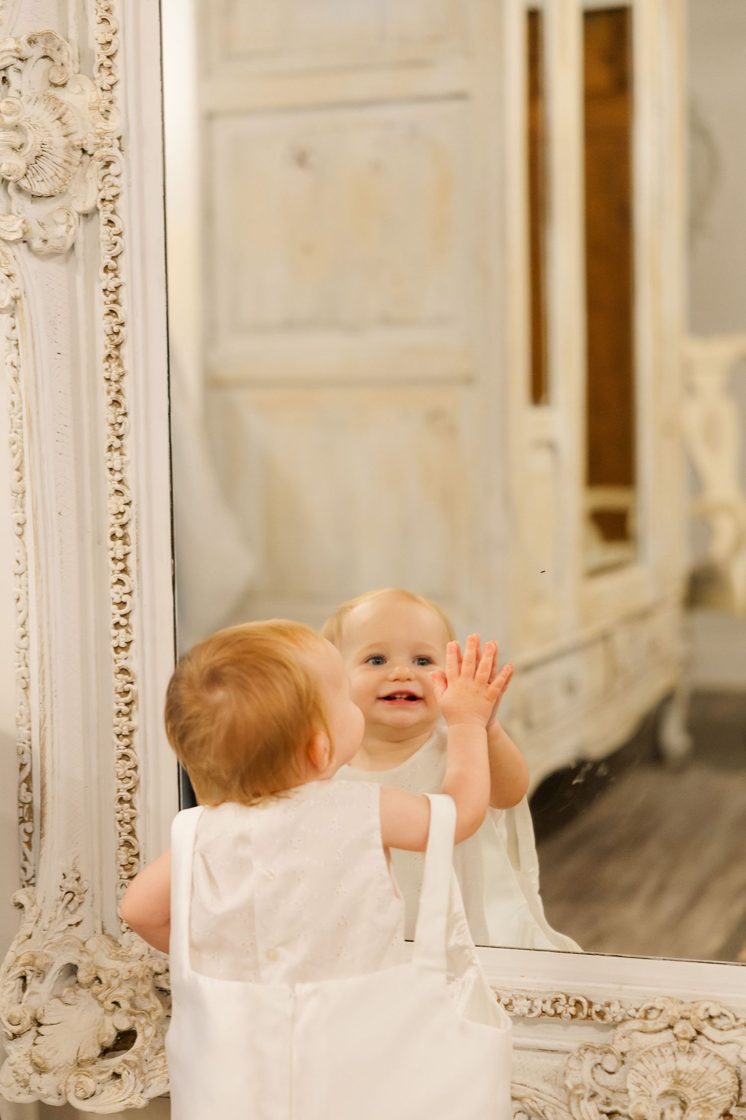 A small child in white dress touches a reflection in an ornate vintage mirror with decorative frame.