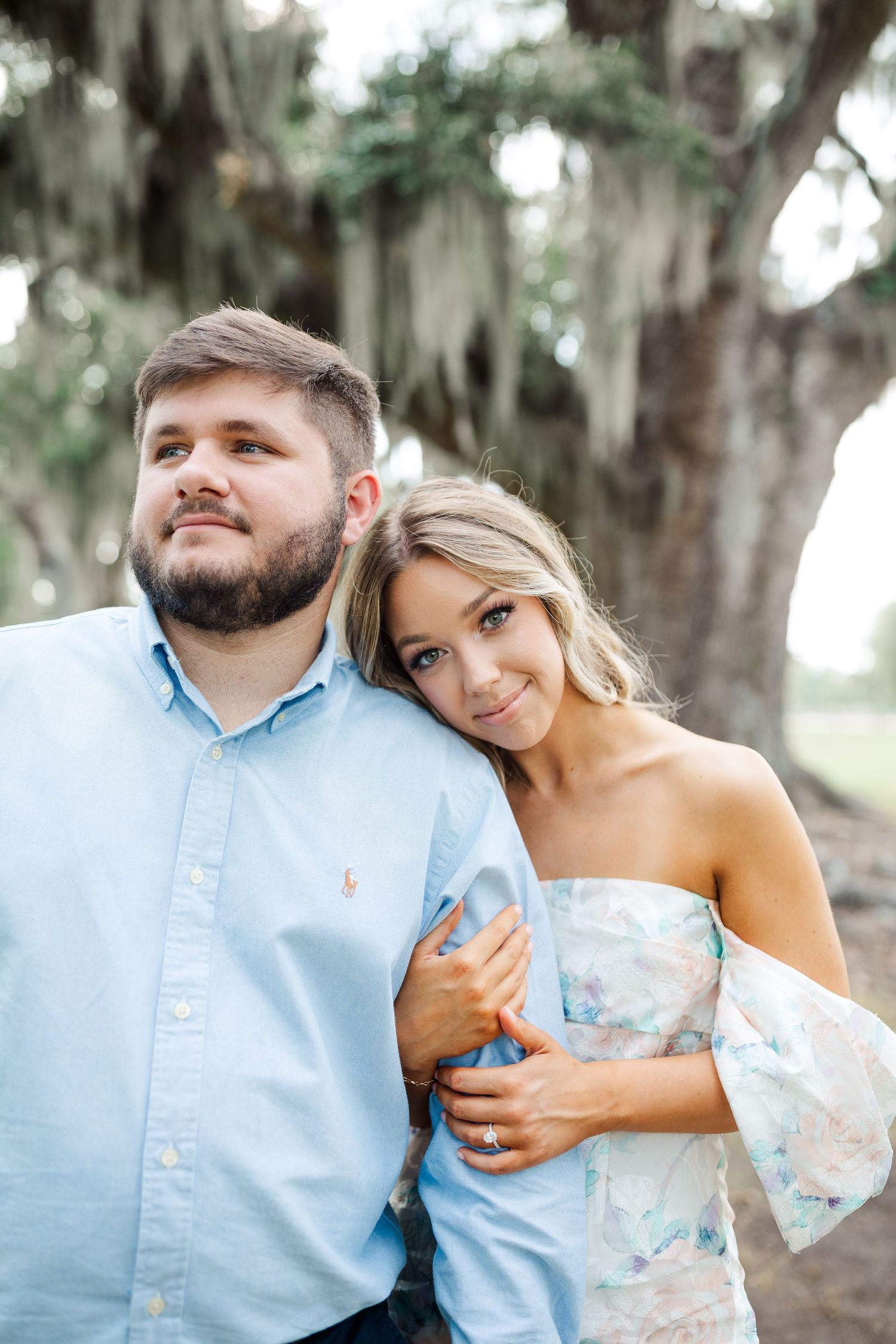 A couple poses together outdoors under Spanish moss trees wearing coordinating light blue outfits.