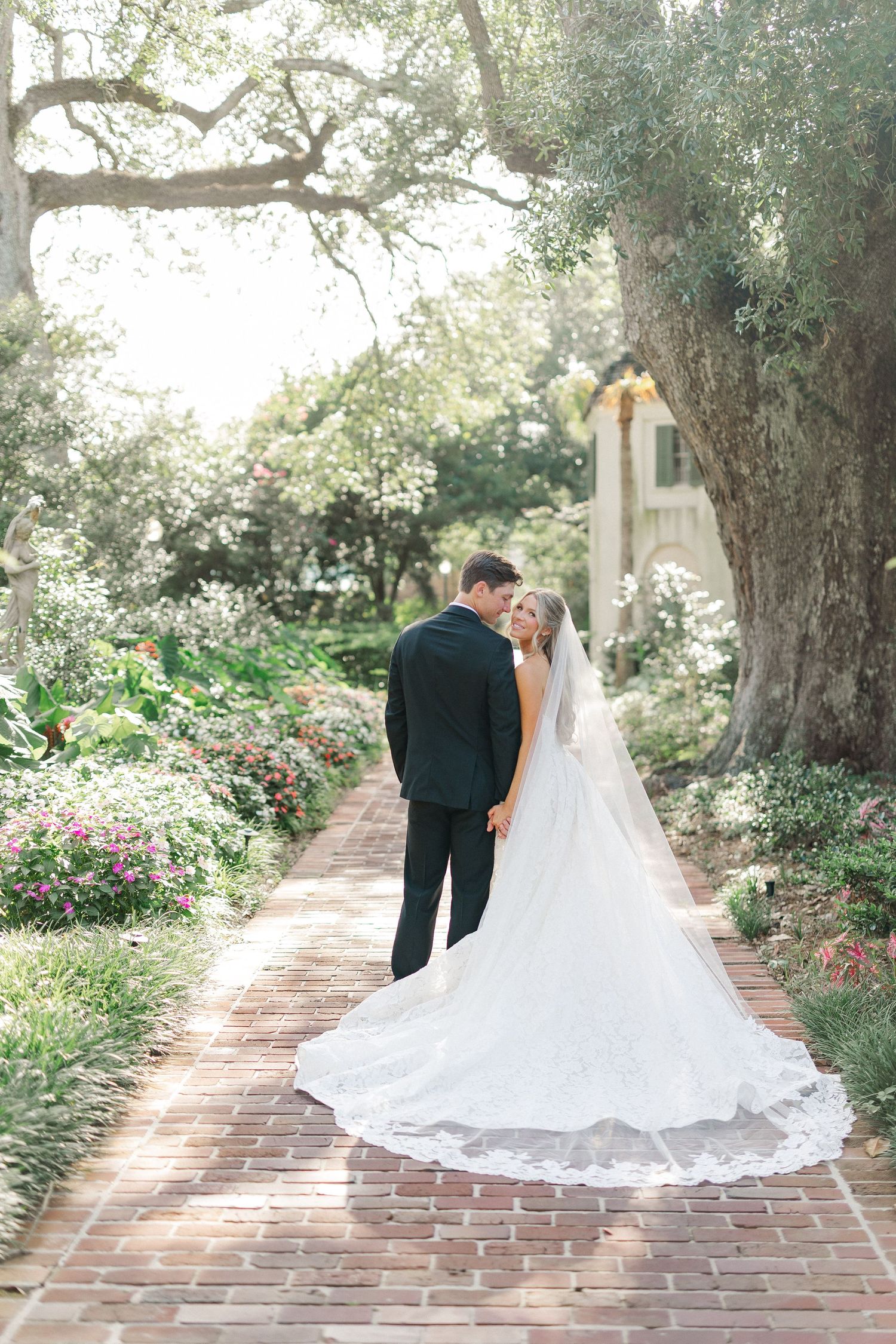Bride and groom share a romantic moment on a brick garden path surrounded by lush greenery and blooming flowers.