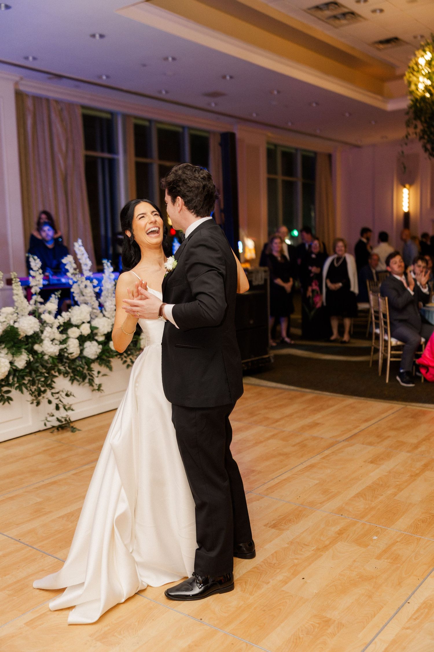 Bride in white gown laughs joyfully while sharing first dance with groom in black tuxedo at wedding reception.