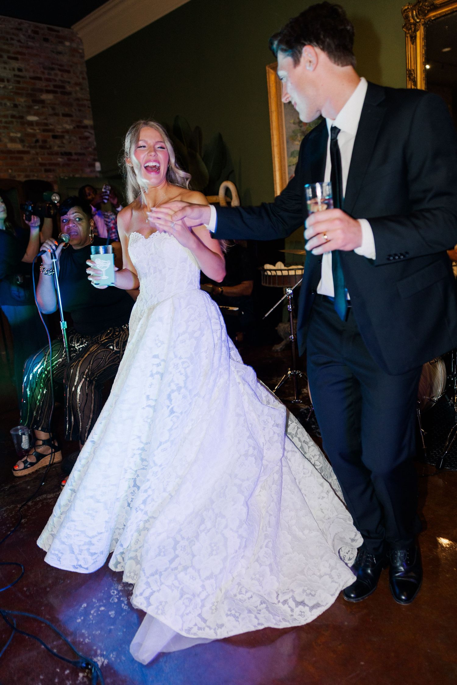 Joyful bride in lace ballgown dances with groom at lively indoor wedding reception with live band.