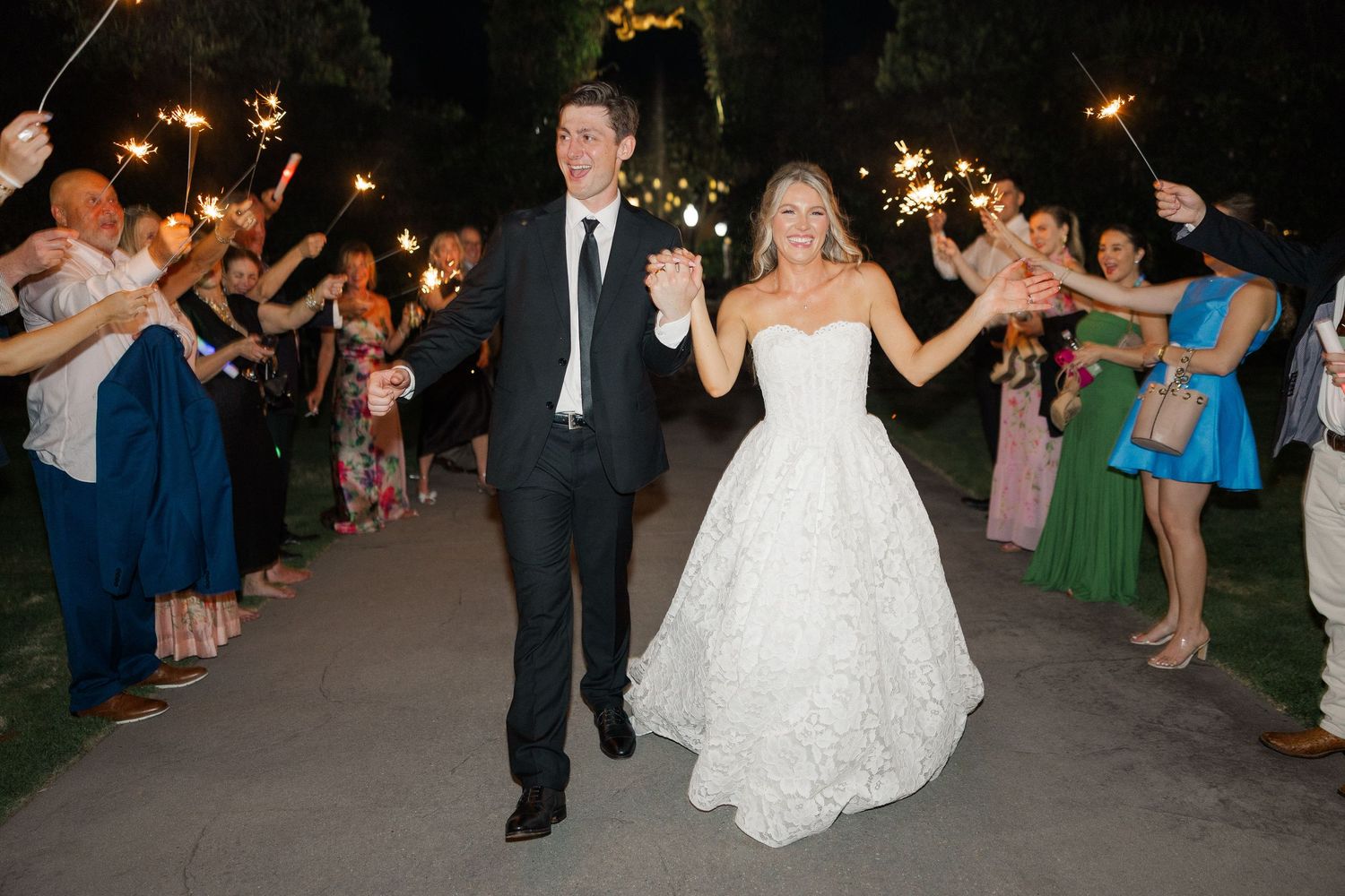 Bride and groom exit wedding reception through sparkler tunnel as smiling guests line the path at night.