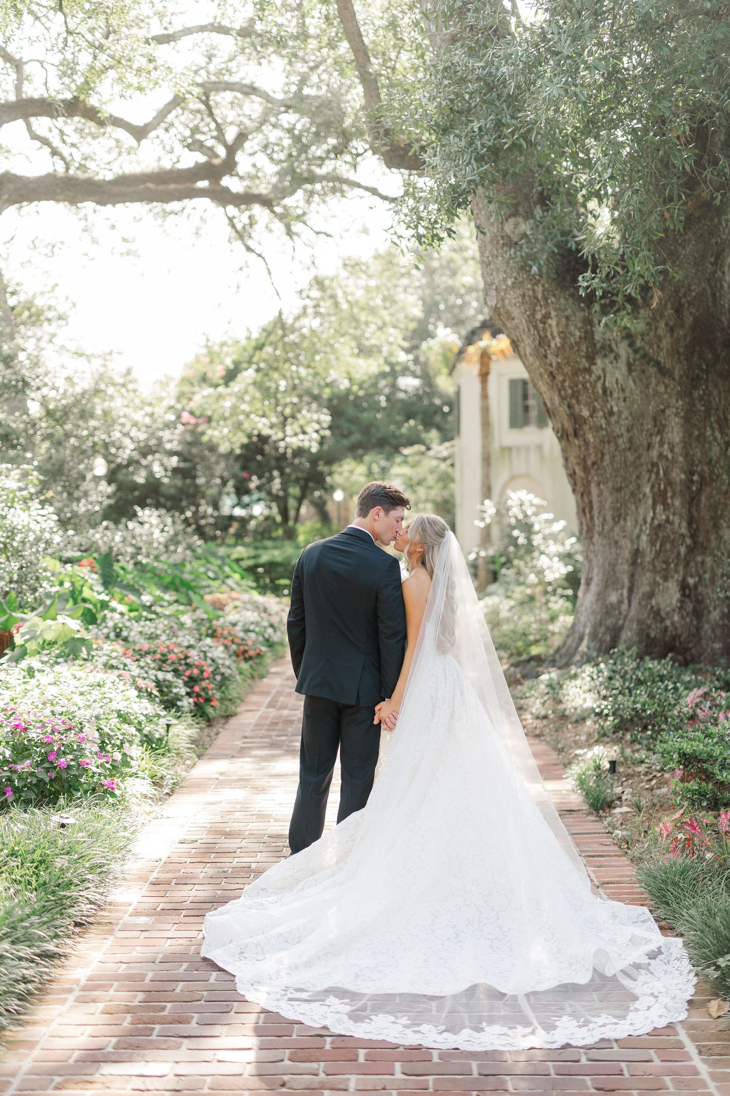 Bride and groom share a romantic kiss on a brick garden path lined with colorful flowers beneath a large oak tree.
