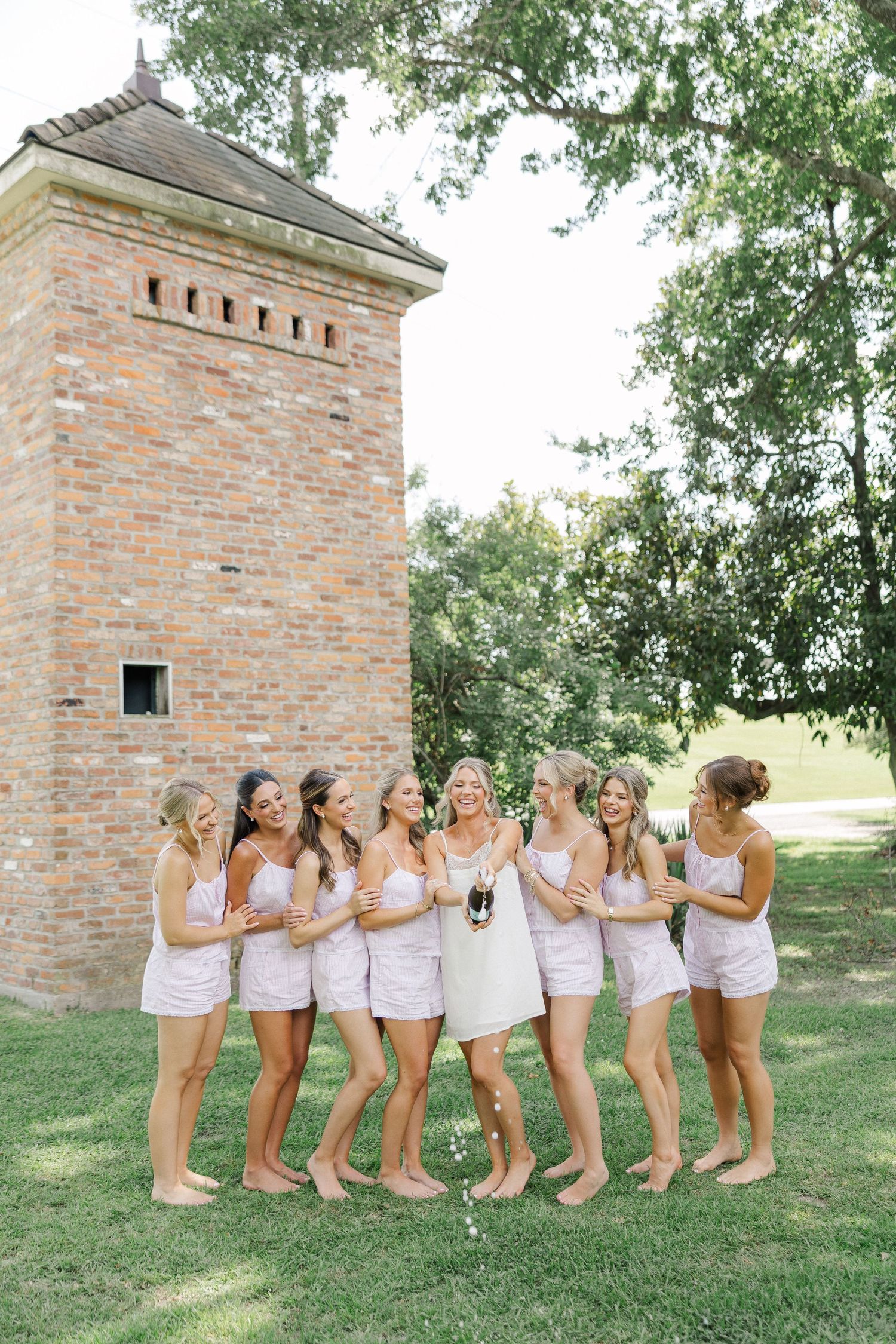 Bridal party in pink matching sets laughing together near rustic brick tower surrounded by trees.