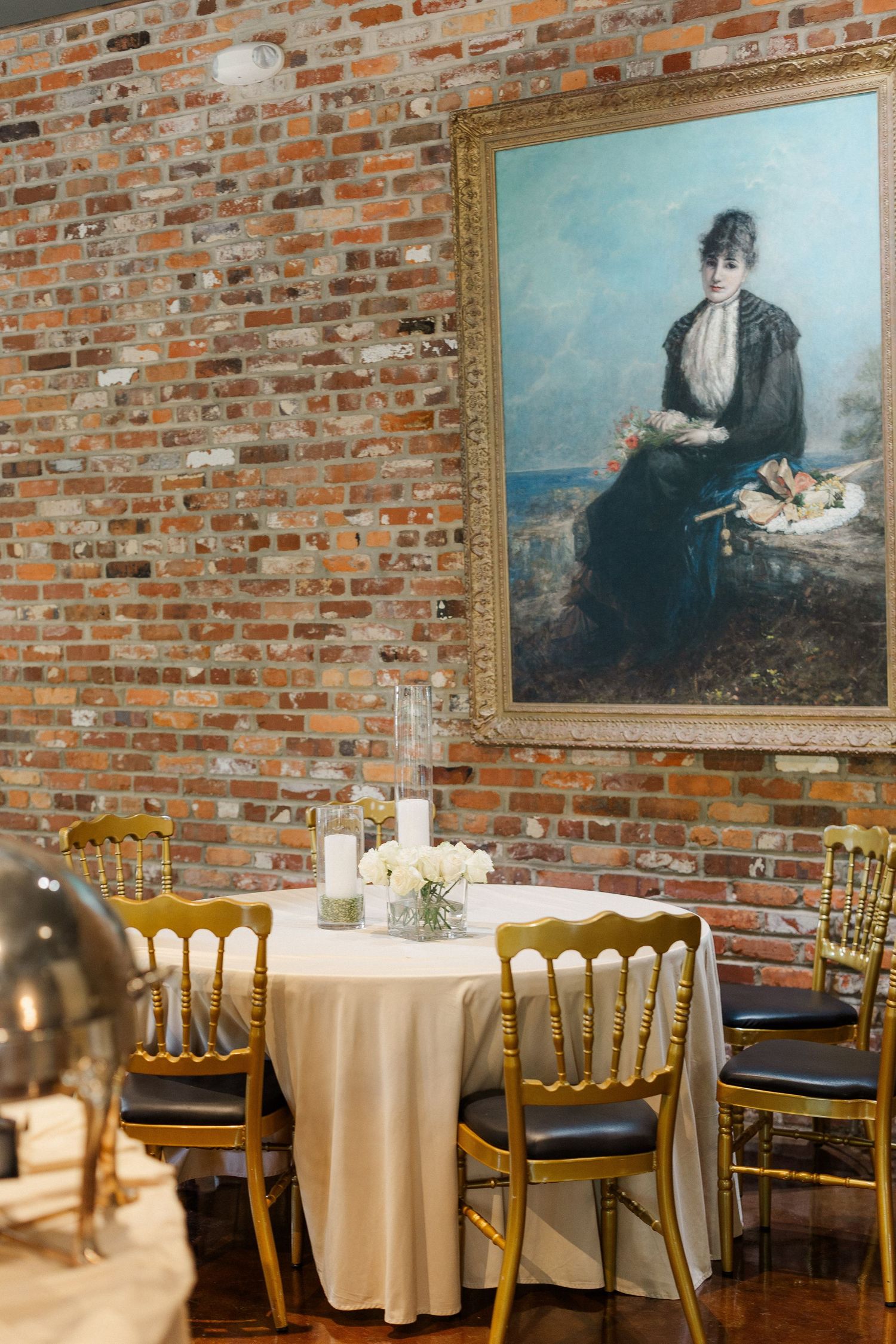 Elegant wedding reception table with gold chairs against exposed brick wall with ornate portrait painting.