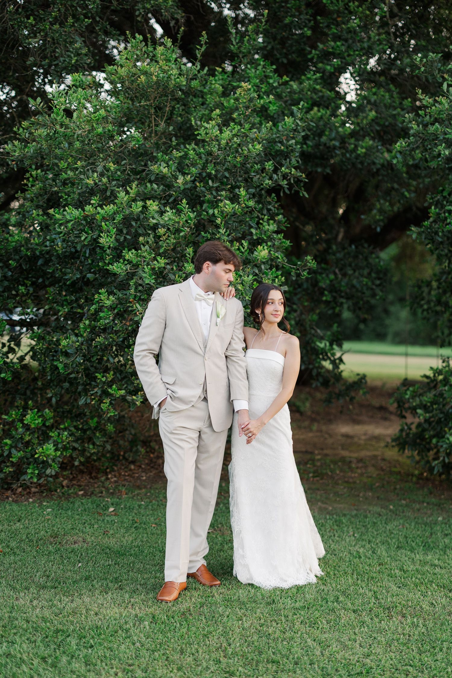 Bride and groom posing together on green lawn in front of lush dark green foliage.