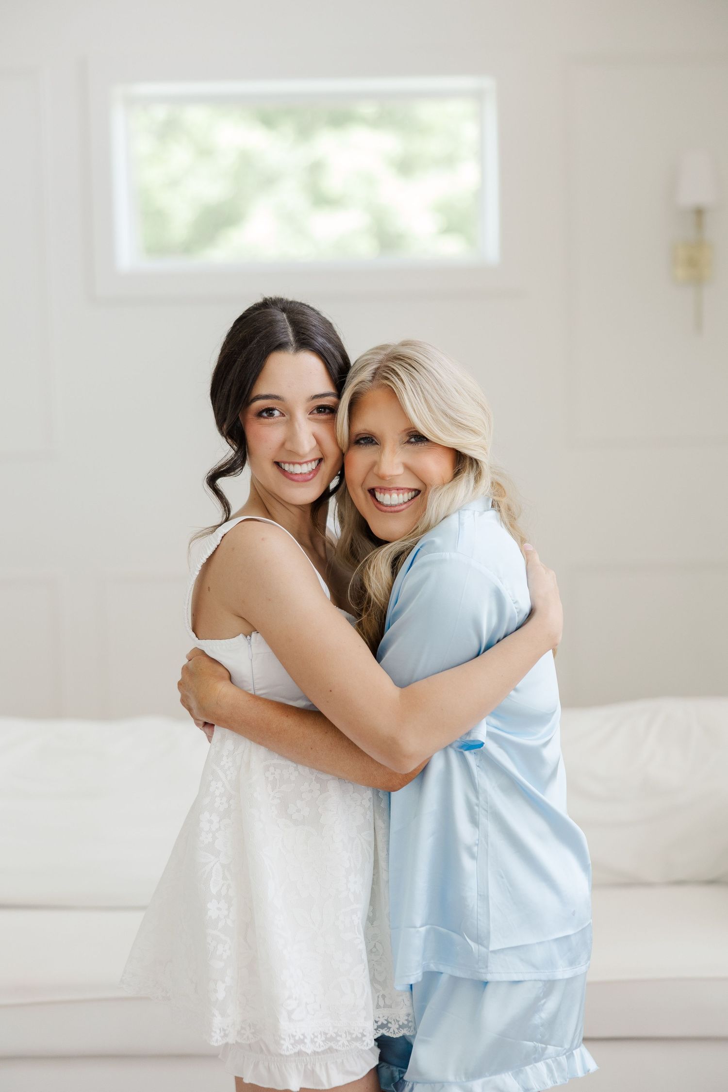 Bride in white dress hugging a woman in blue satin pajamas, smiling together in a bright white room.