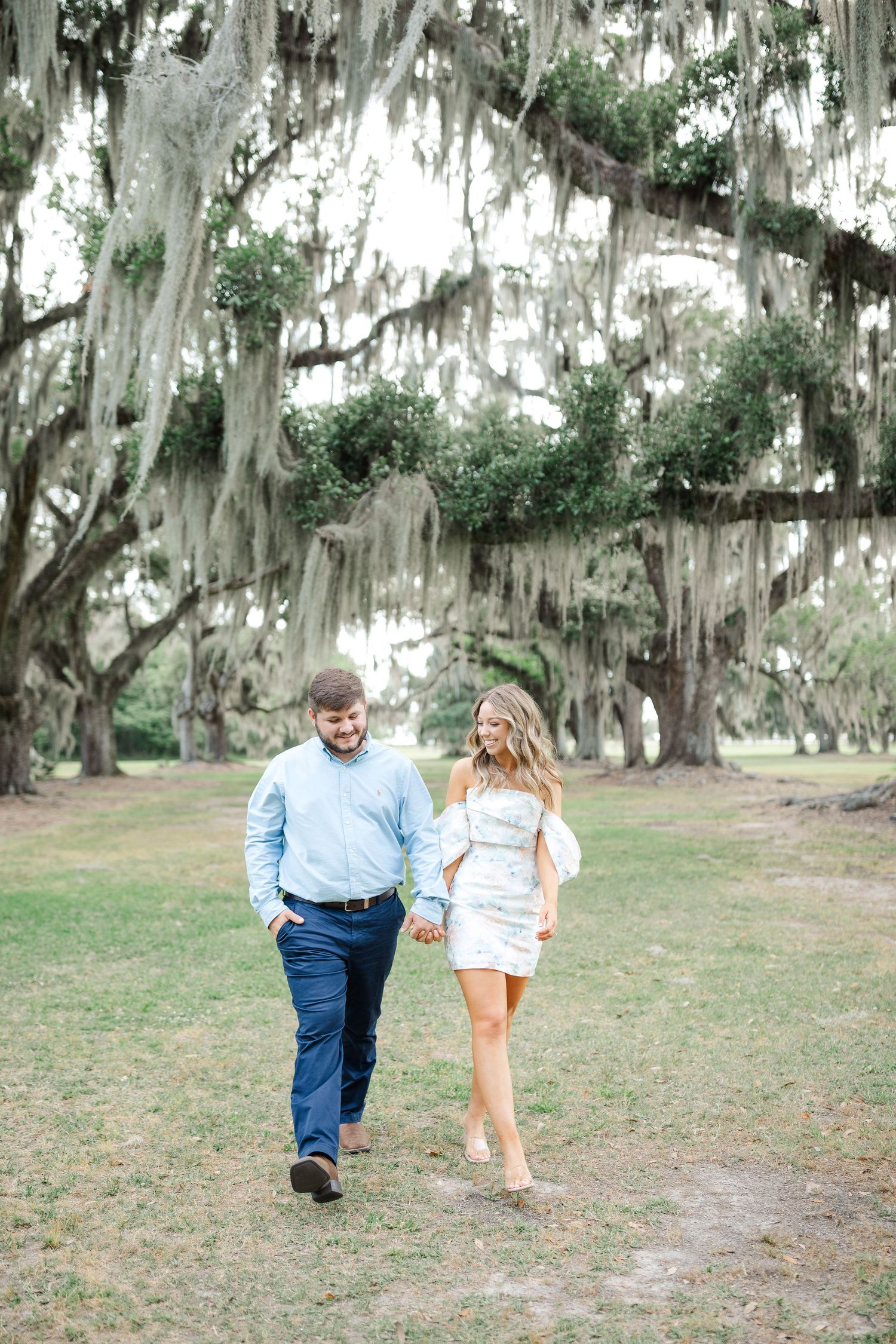 A couple strolls hand in hand beneath Spanish moss-draped oak trees in a Southern park setting.