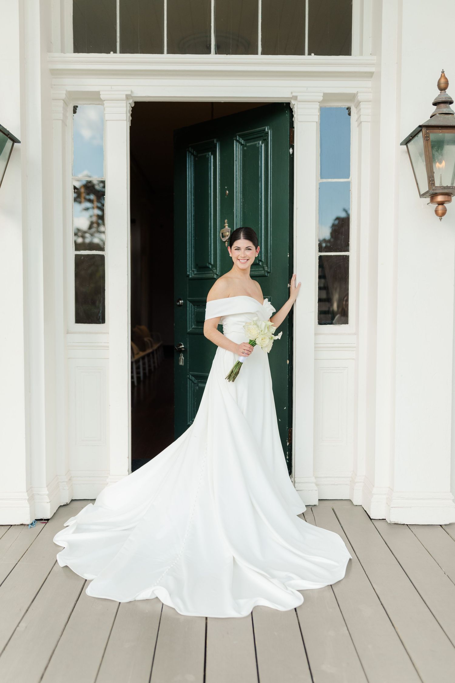 An elegant wedding dress with a flowing train is showcased on a white columned porch with a dark green door.