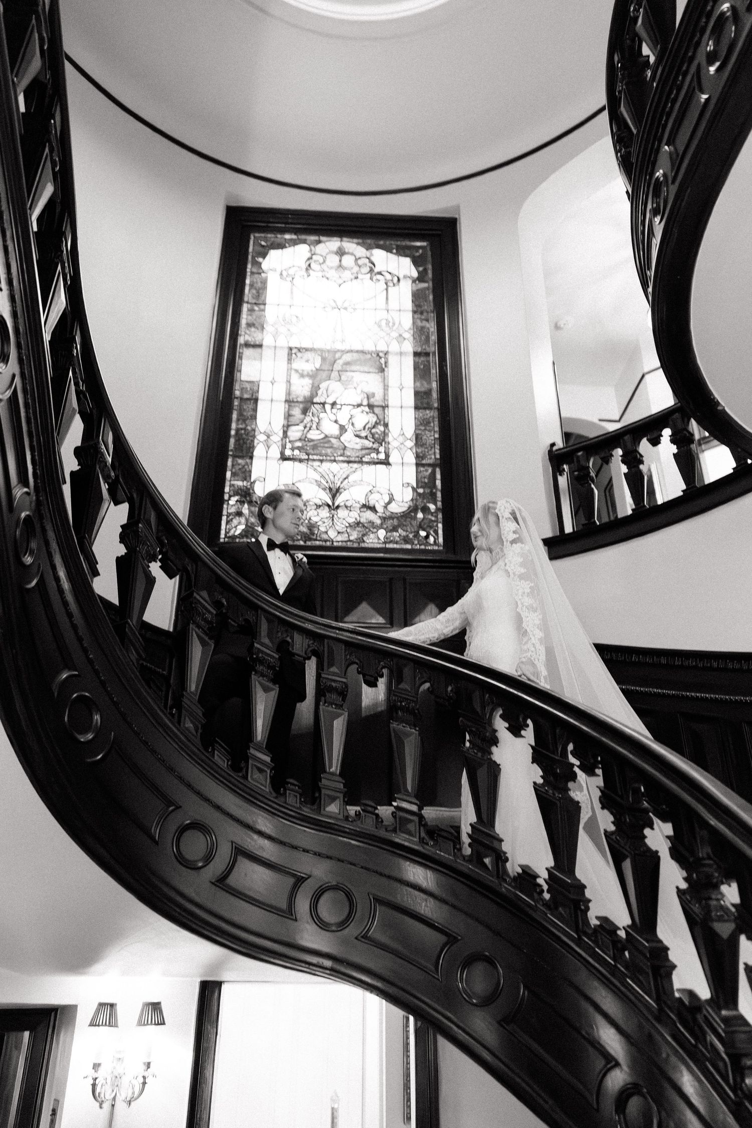 Black and white photo of an ornate staircase with decorative railings and stained glass windows.