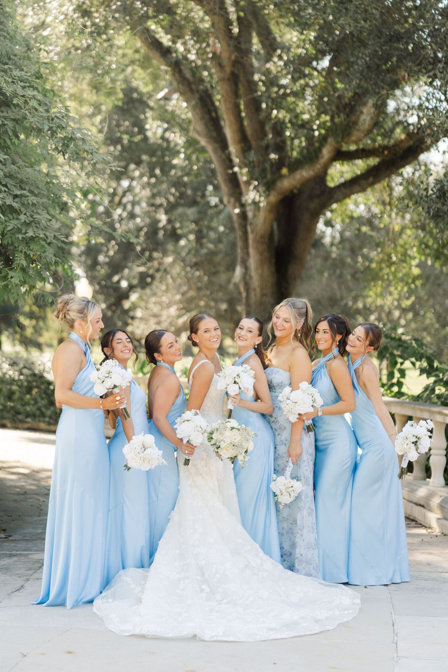 Bridal party in powder blue dresses and white gown pose together under trees for outdoor wedding photos.