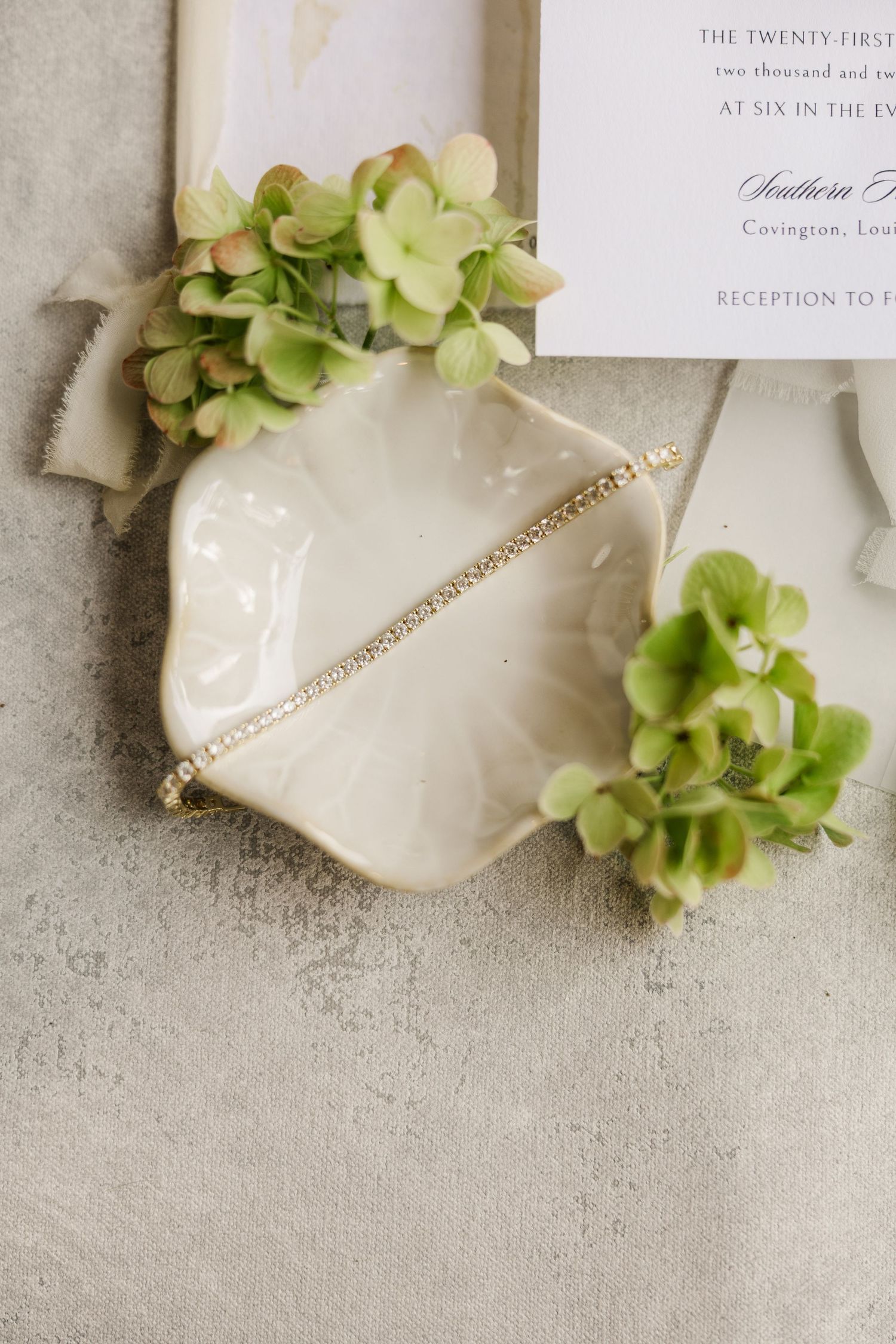 Wedding details flatlay featuring white paper invitations, green floral accents, and pearl jewelry on a neutral background.