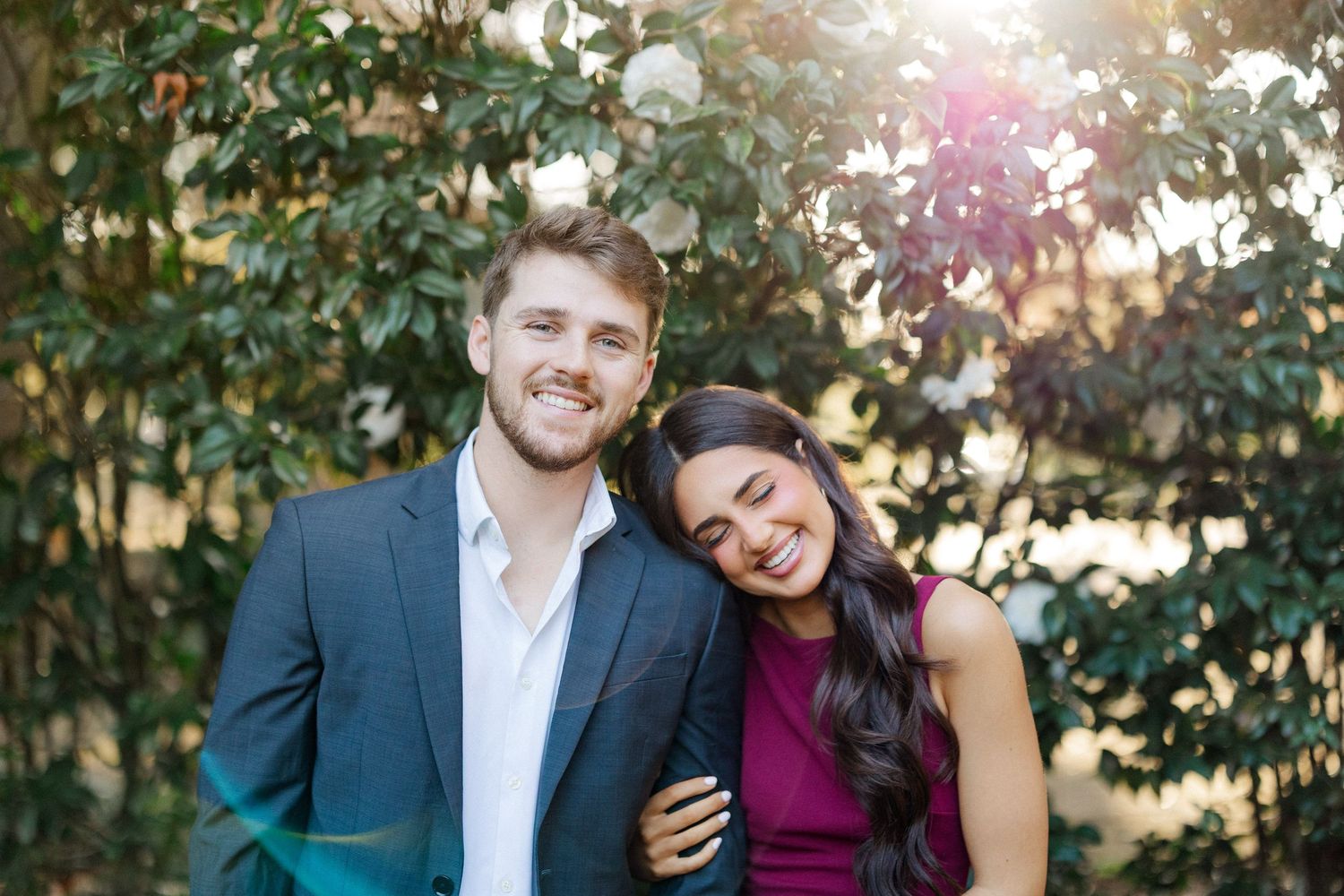 A couple laughs together during an outdoor engagement photo session with greenery in the background.
