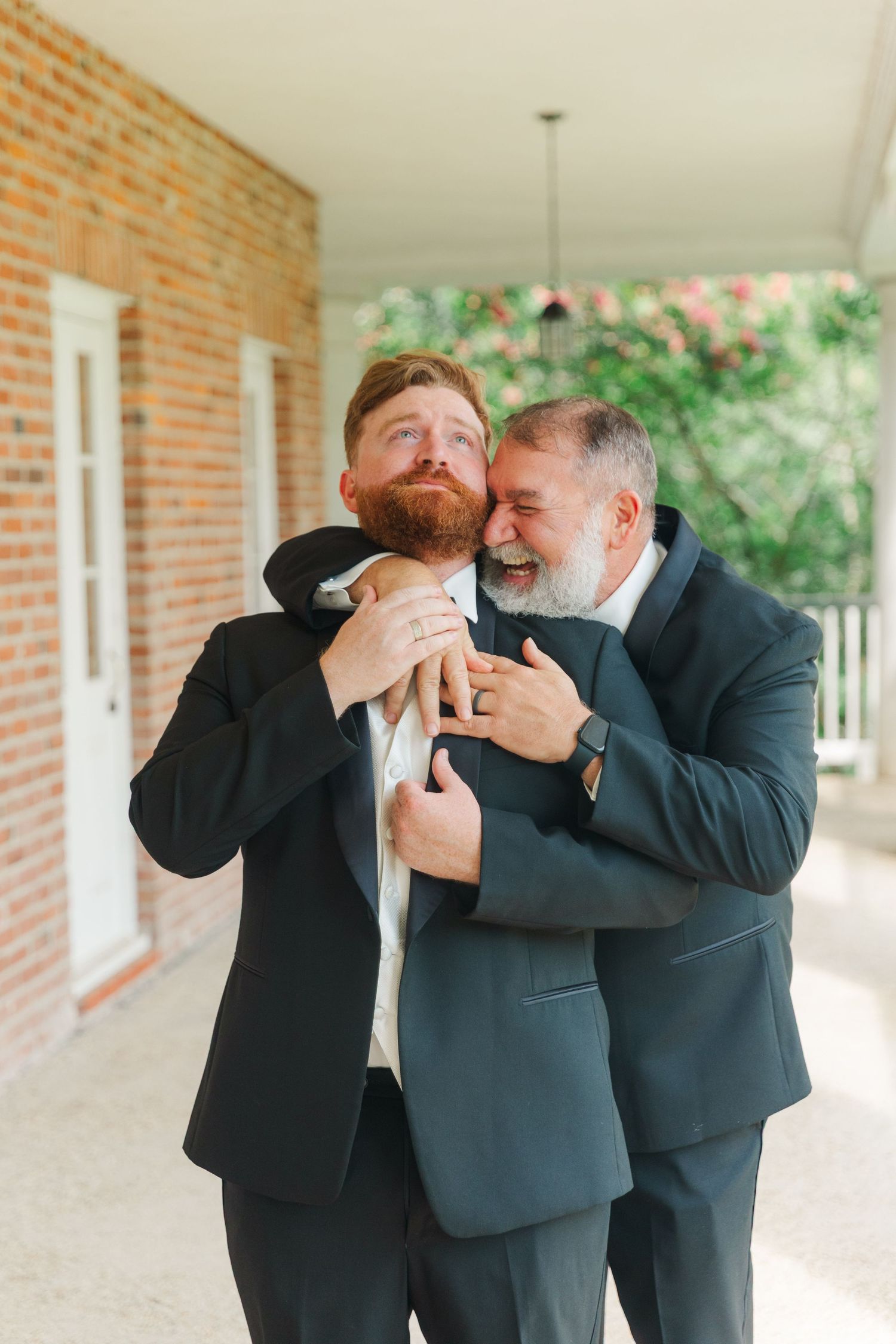 Groomsmen share a lighthearted moment laughing together in formal suits against a brick wall backdrop.