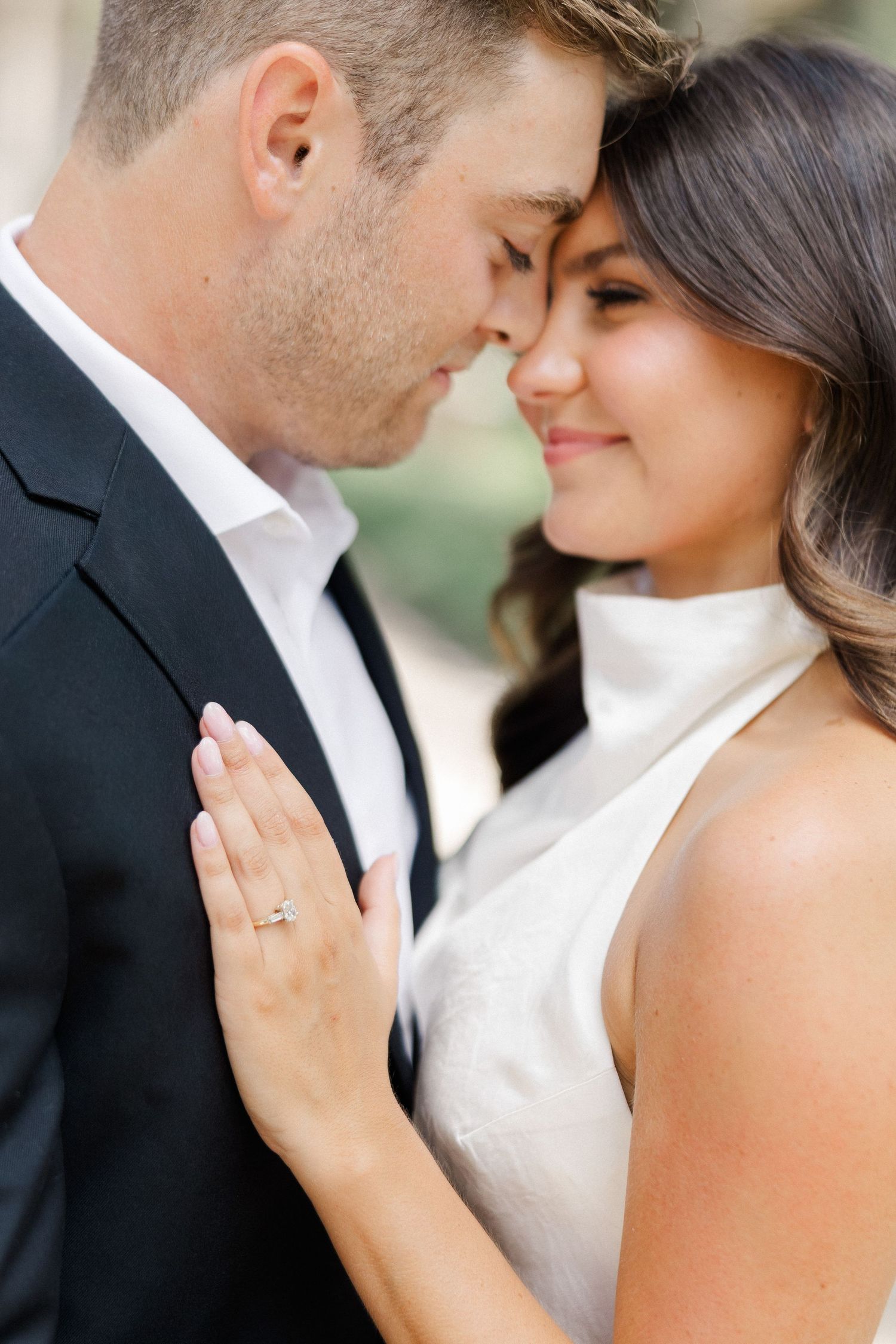A couple in elegant attire share an intimate moment, the woman smiling softly while wearing a diamond engagement ring.