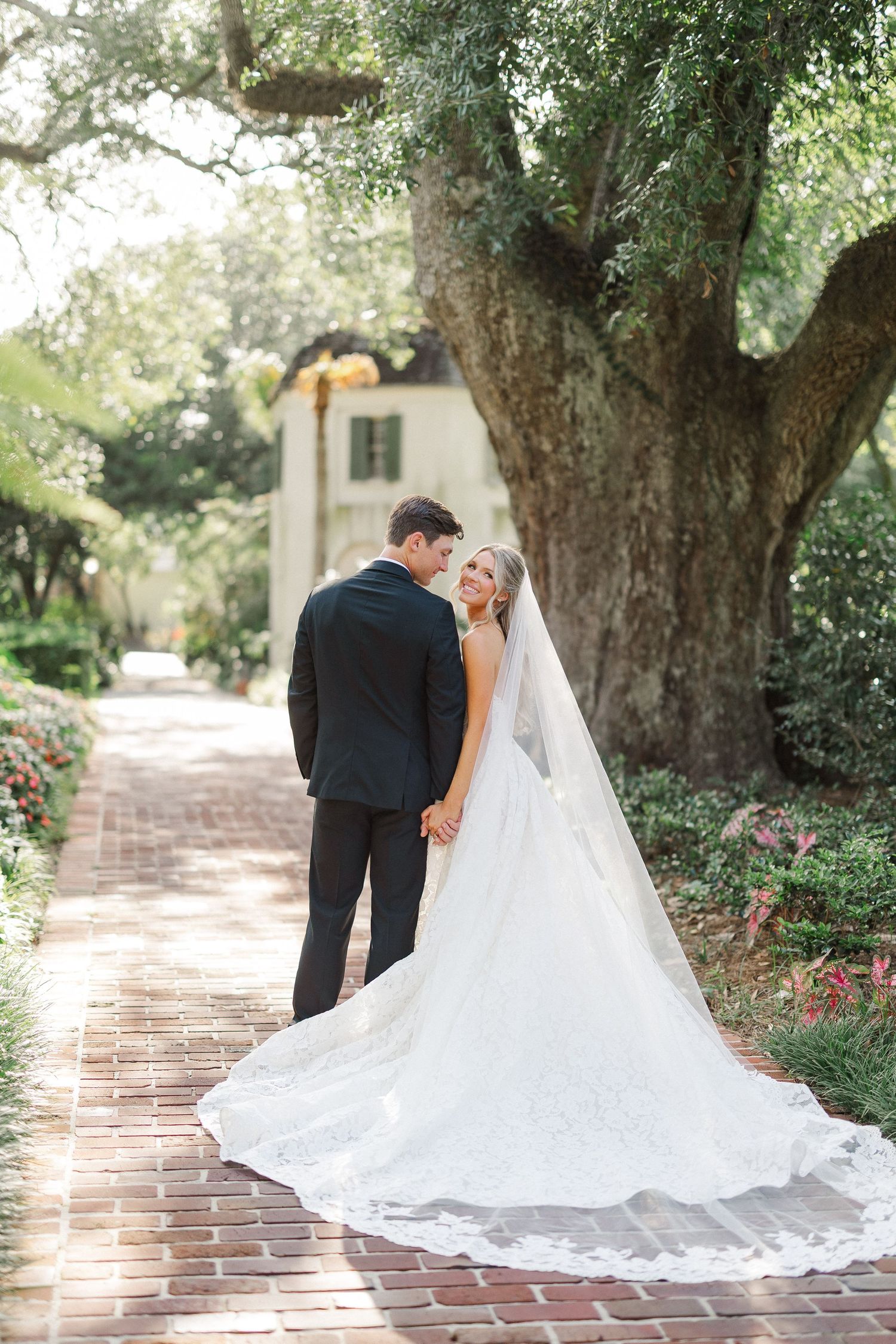 Bride in lace cathedral veil gown and groom in black suit on brick path under oak tree.