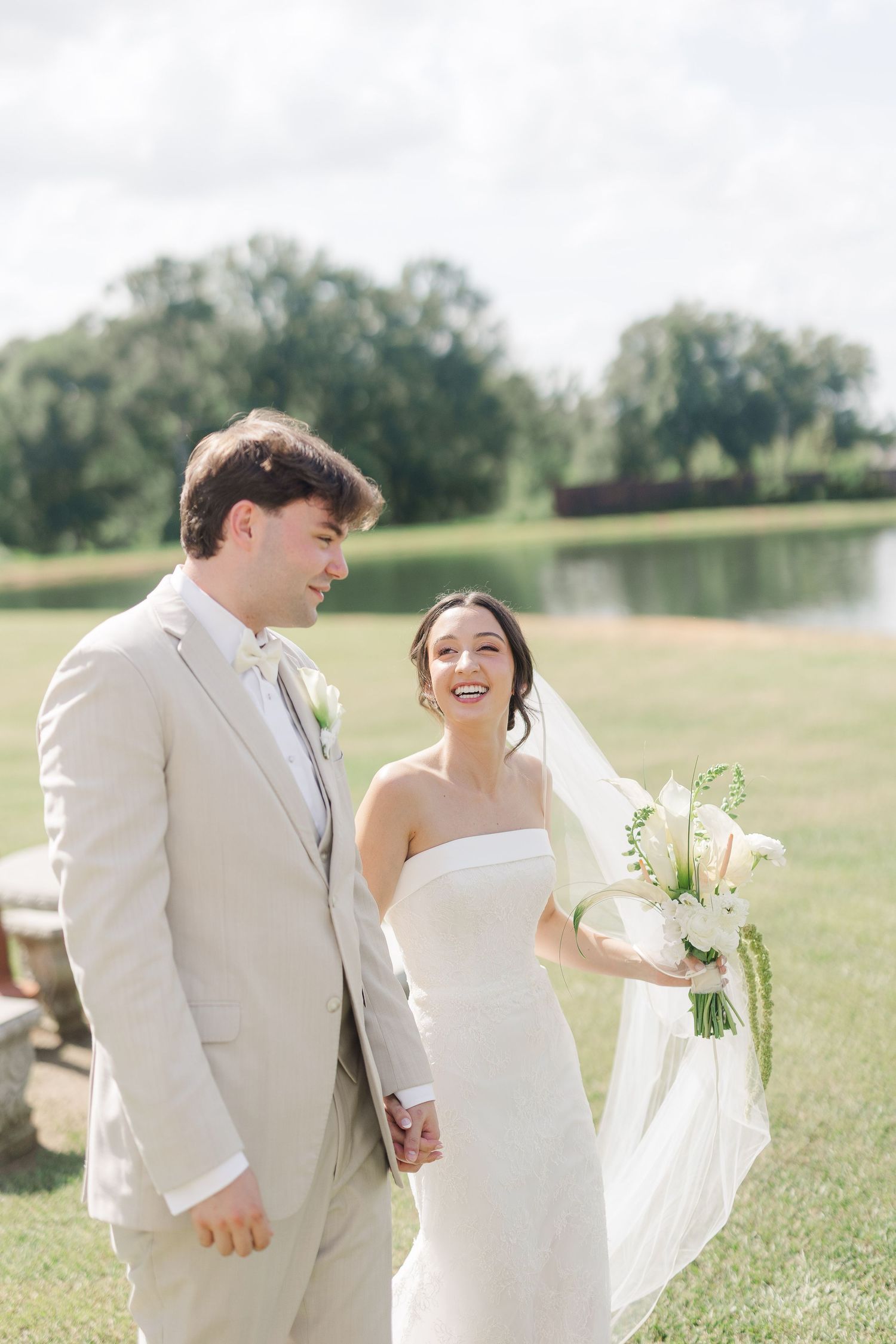 Bride and groom walk hand in hand outdoors, bride holding white bouquet with veil flowing in the breeze.