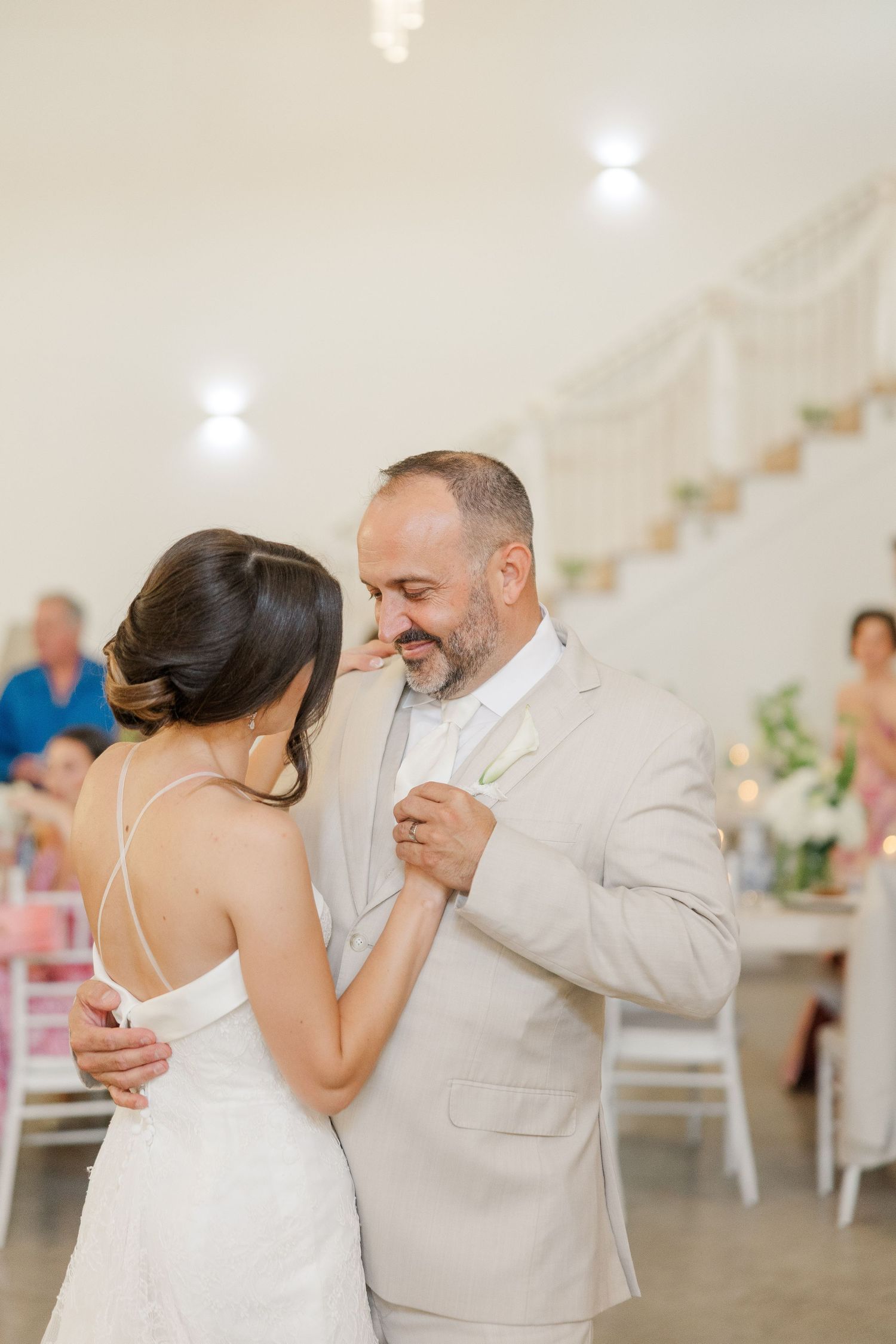 Bride and older man in beige suit share an emotional father-daughter dance at elegant wedding reception.
