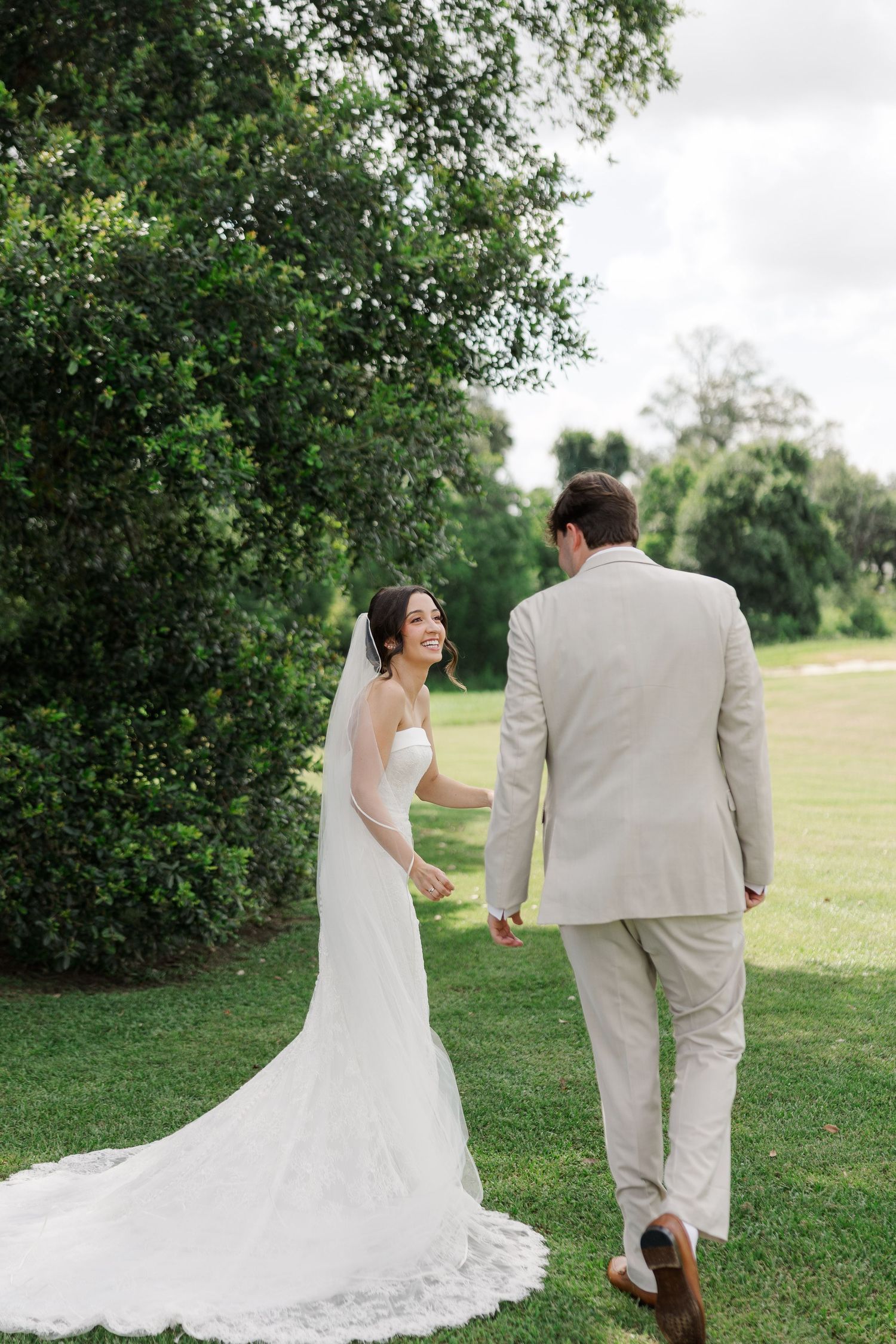 Bride in white gown and veil smiles at groom in tan suit as they walk hand in hand across lush green lawn.