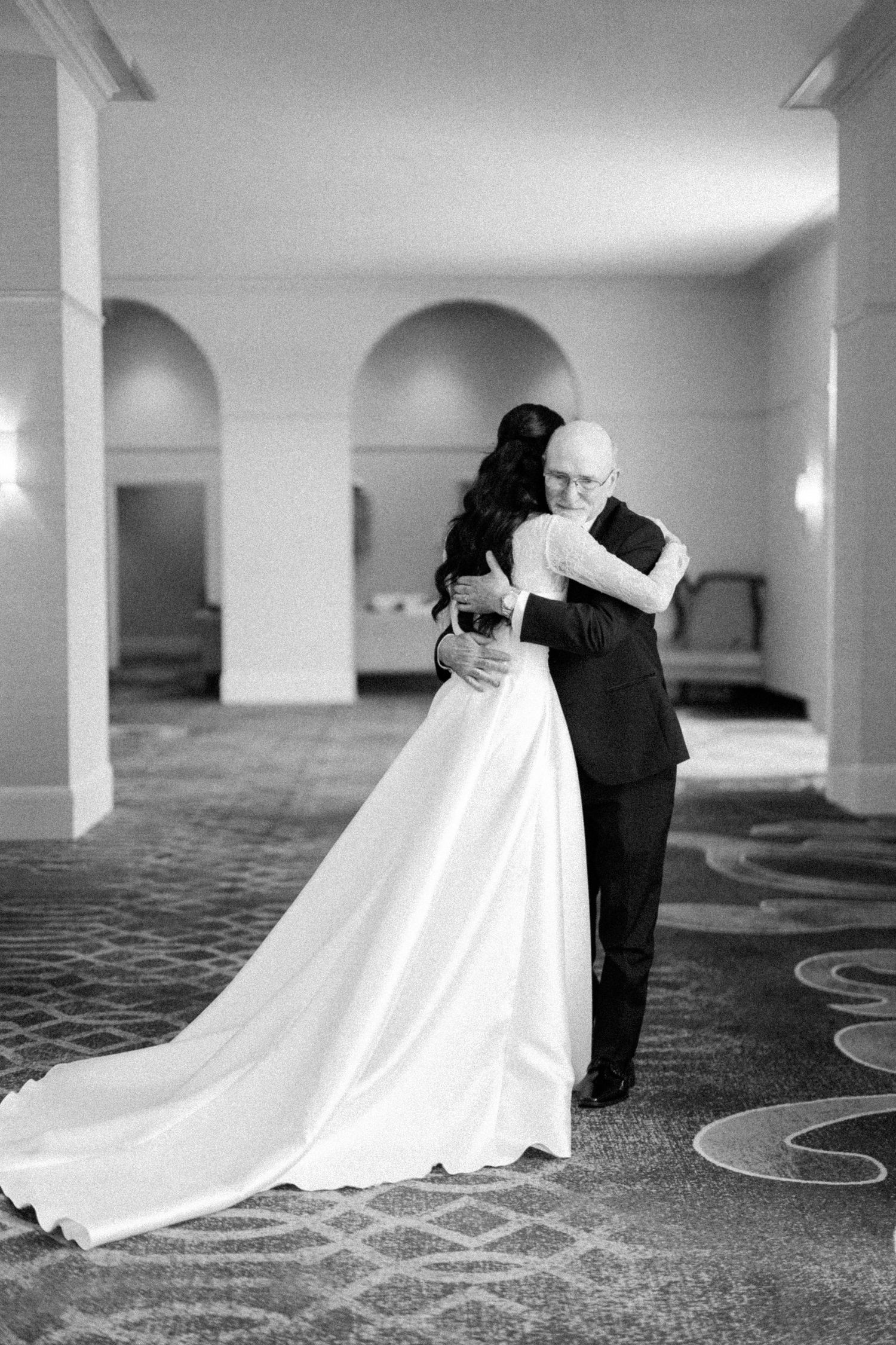 Black and white photo of bride in elegant gown embracing man in tuxedo in grand hall with arched ceilings.