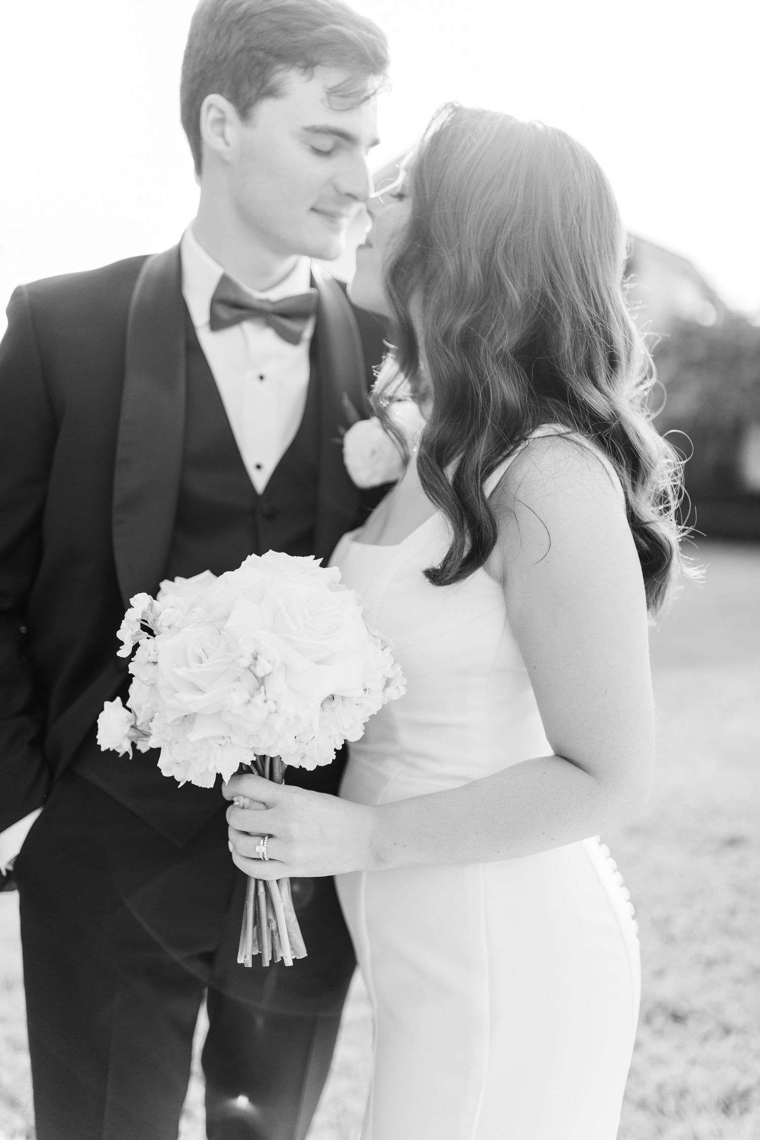 Black and white wedding photo of bride holding white bouquet with groom in tuxedo gazing at each other outdoors.