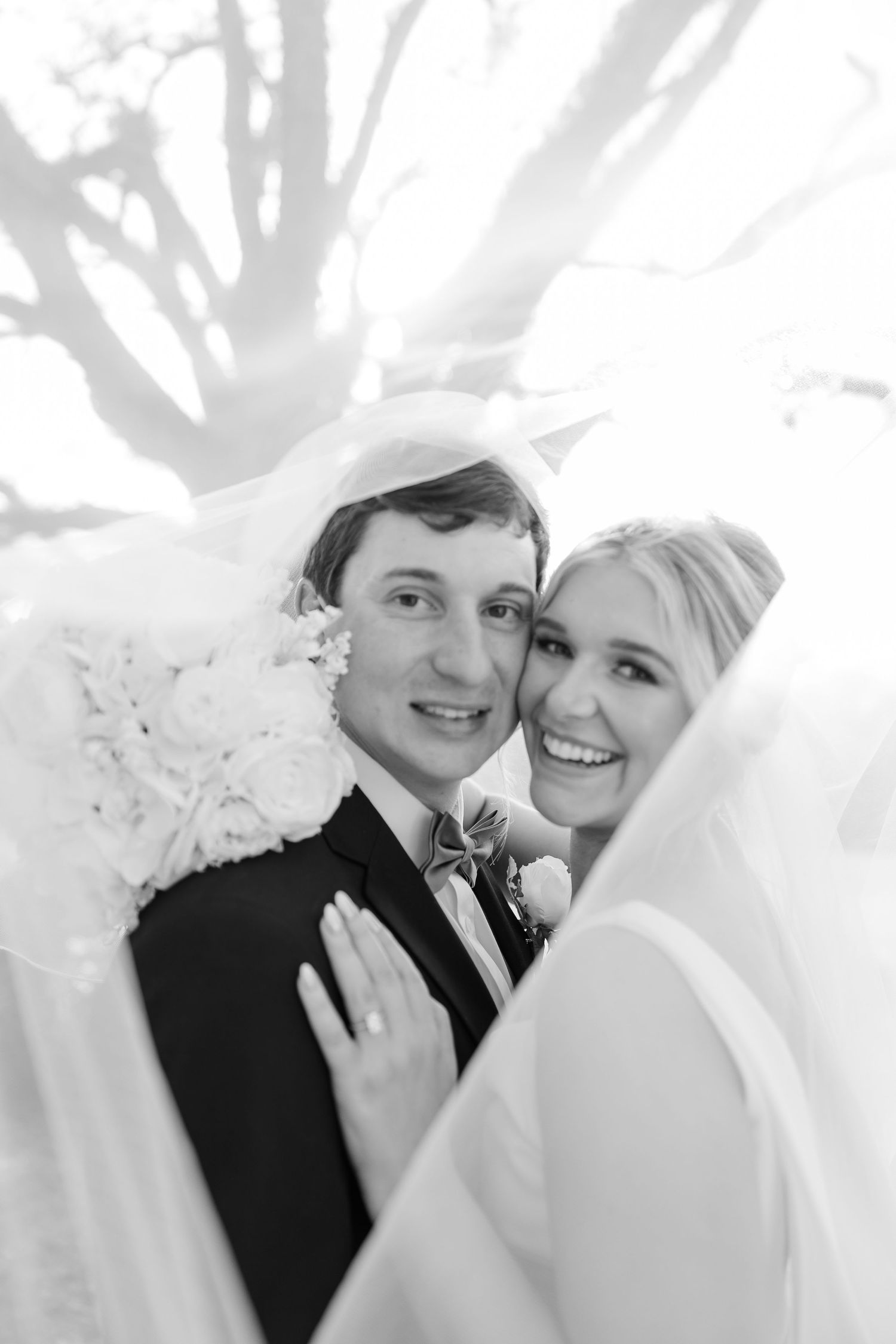 A black and white wedding photograph capturing a romantic moment between a couple under a veil.